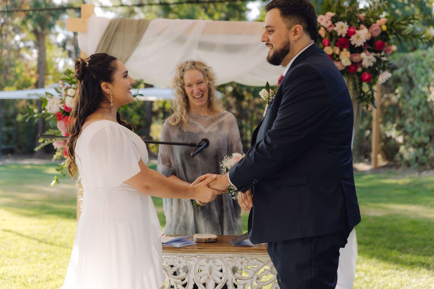 Bride and groom hold hands and smile during outdoor wedding ceremony with blonde officiant behind them.