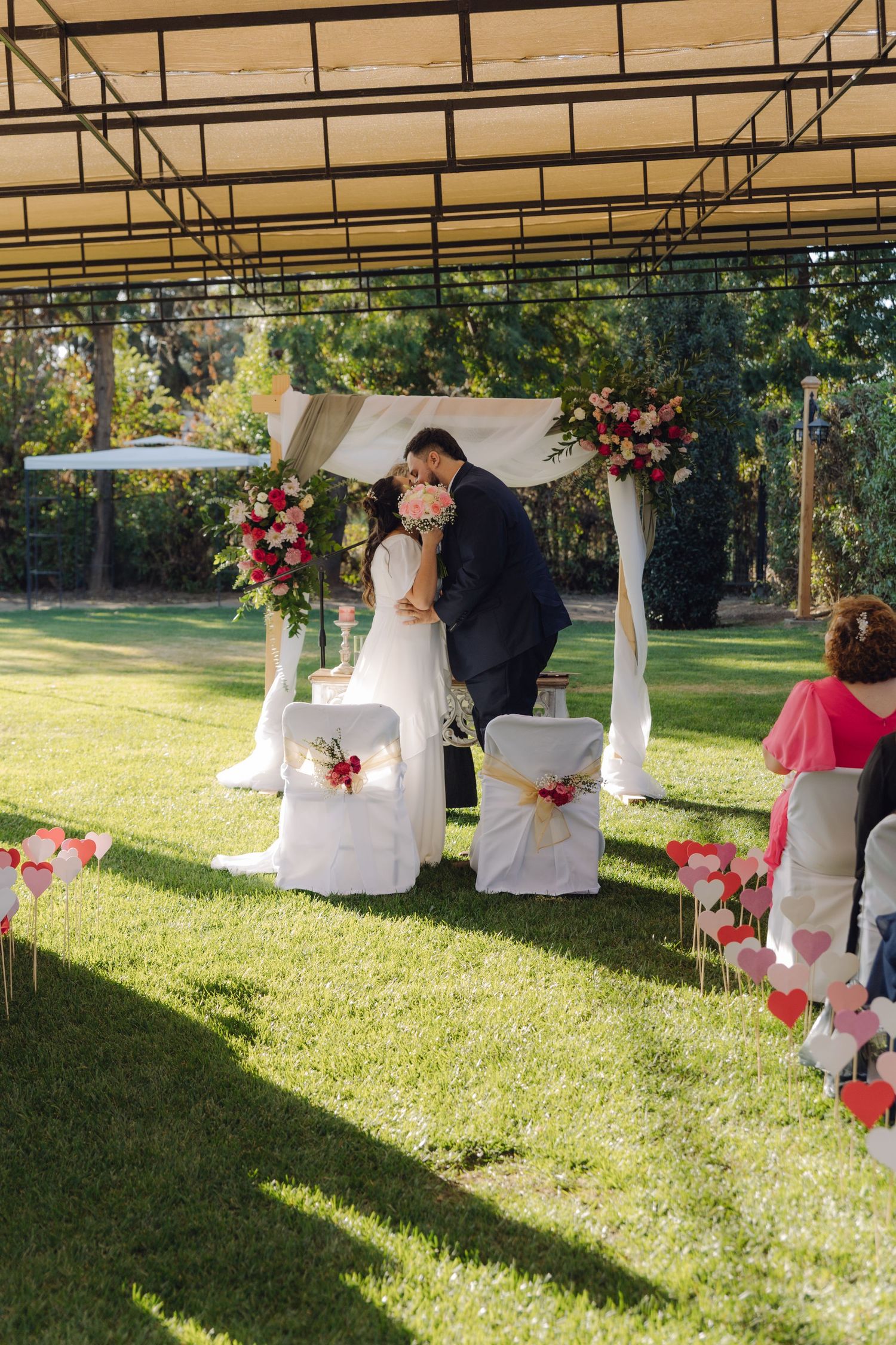 Newlywed couple shares first kiss under floral arch at outdoor garden wedding ceremony.