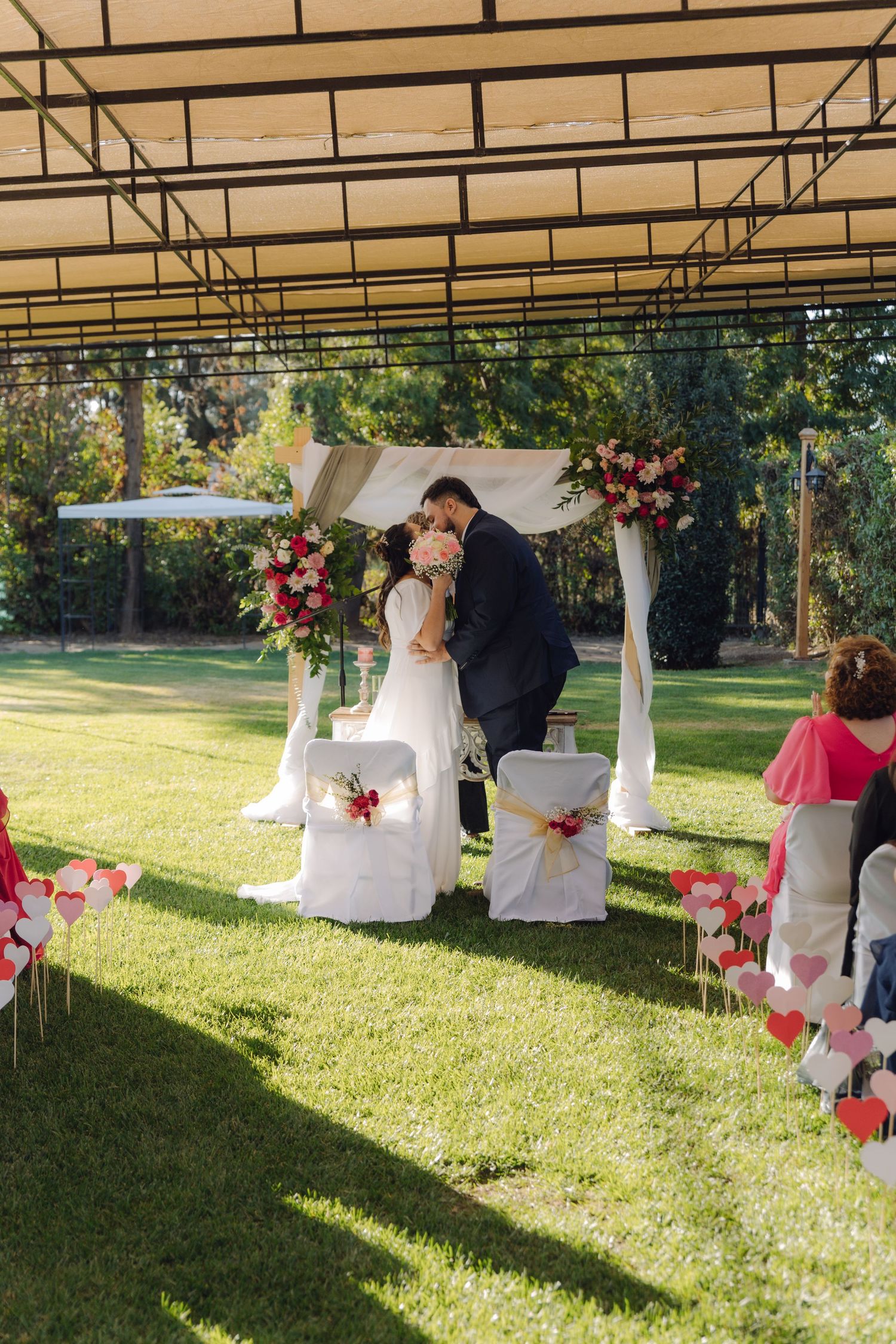 Bride and groom kiss beneath decorated wedding arch surrounded by heart decorations on sunny lawn.