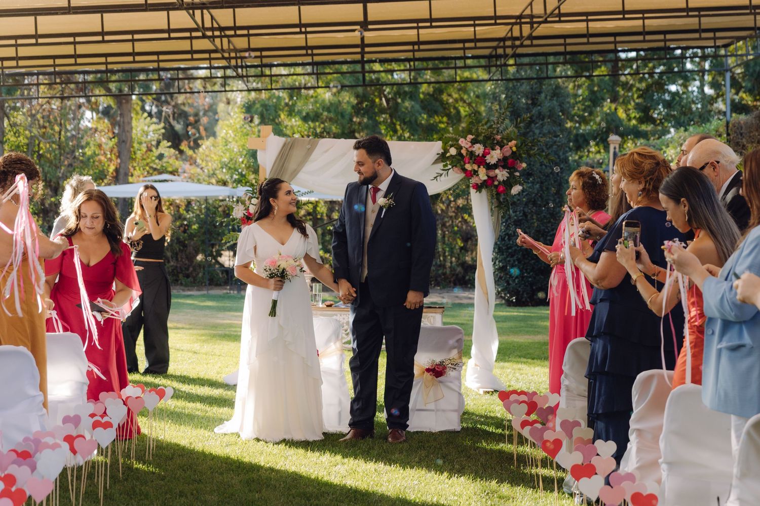 Newlyweds walk down aisle as guests celebrate with pink ribbons at outdoor garden wedding ceremony.