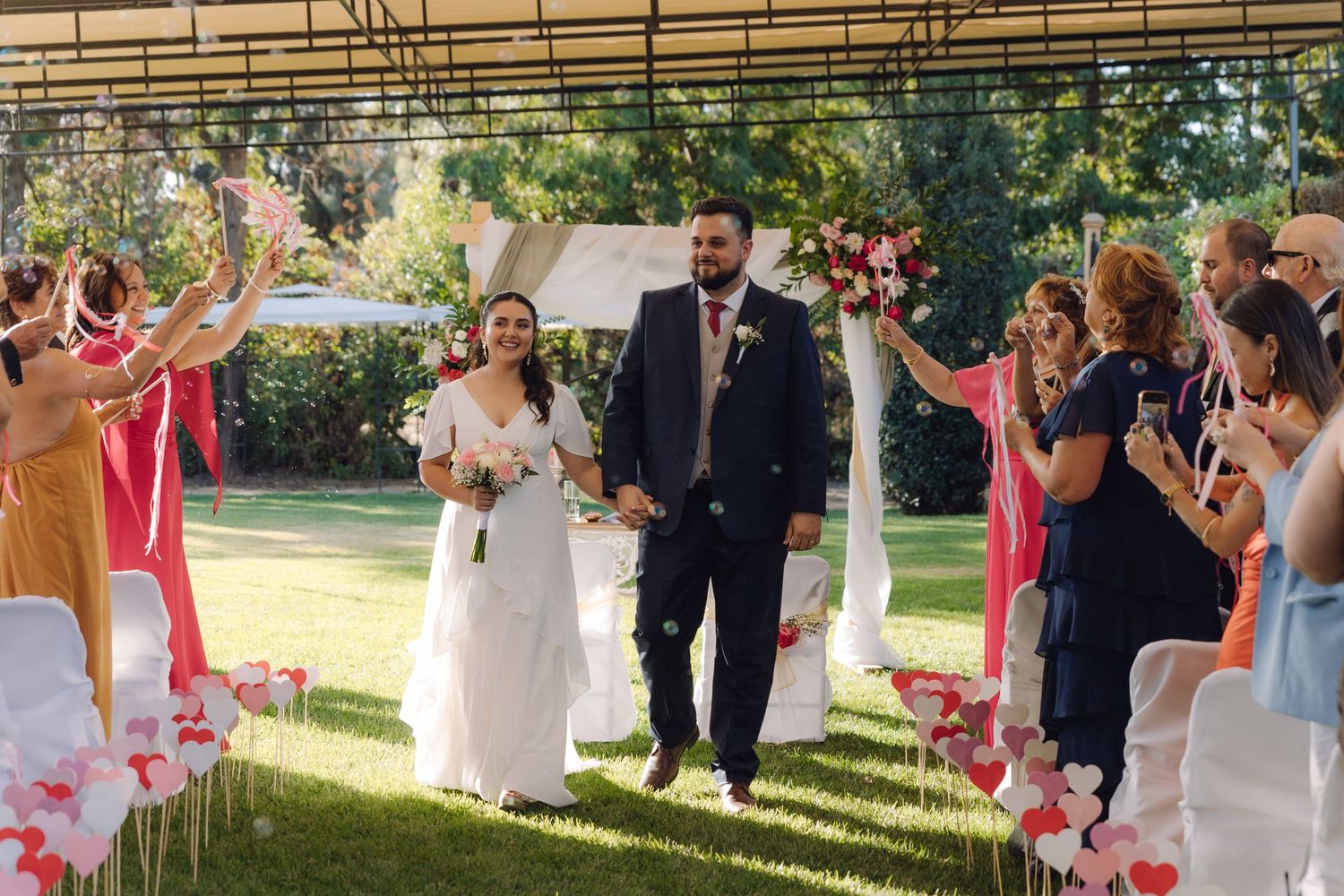 Smiling newlyweds walk down aisle as guests toss confetti at outdoor garden wedding ceremony.