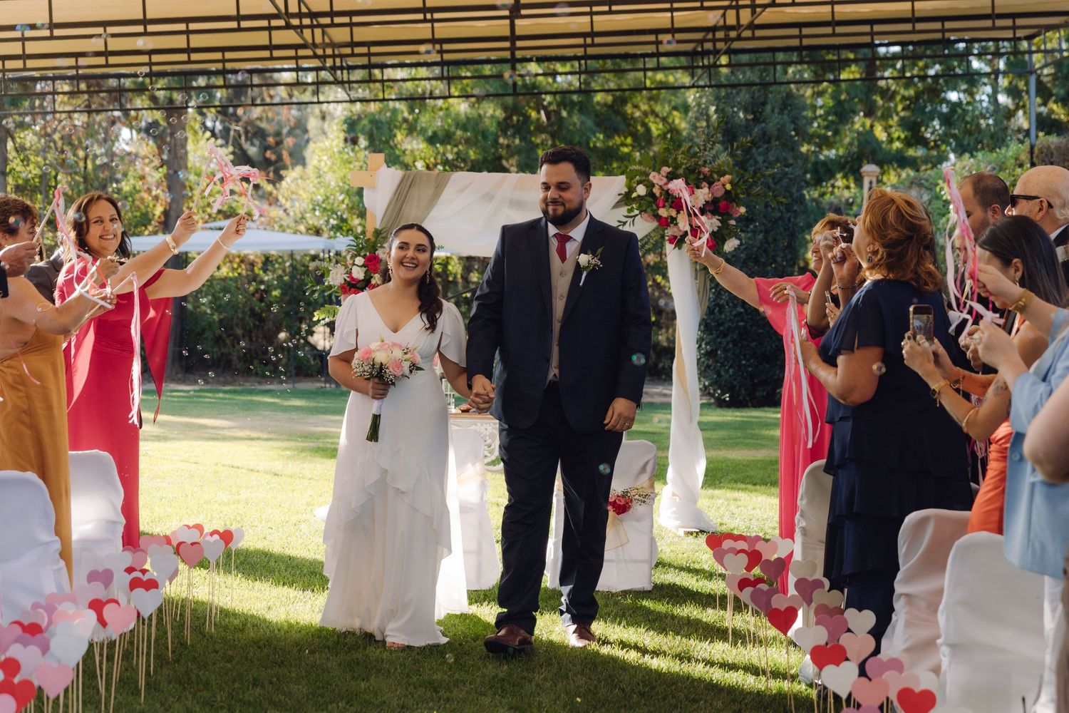 Joyful newlyweds walk through cheering guests after outdoor garden wedding ceremony on sunny day.