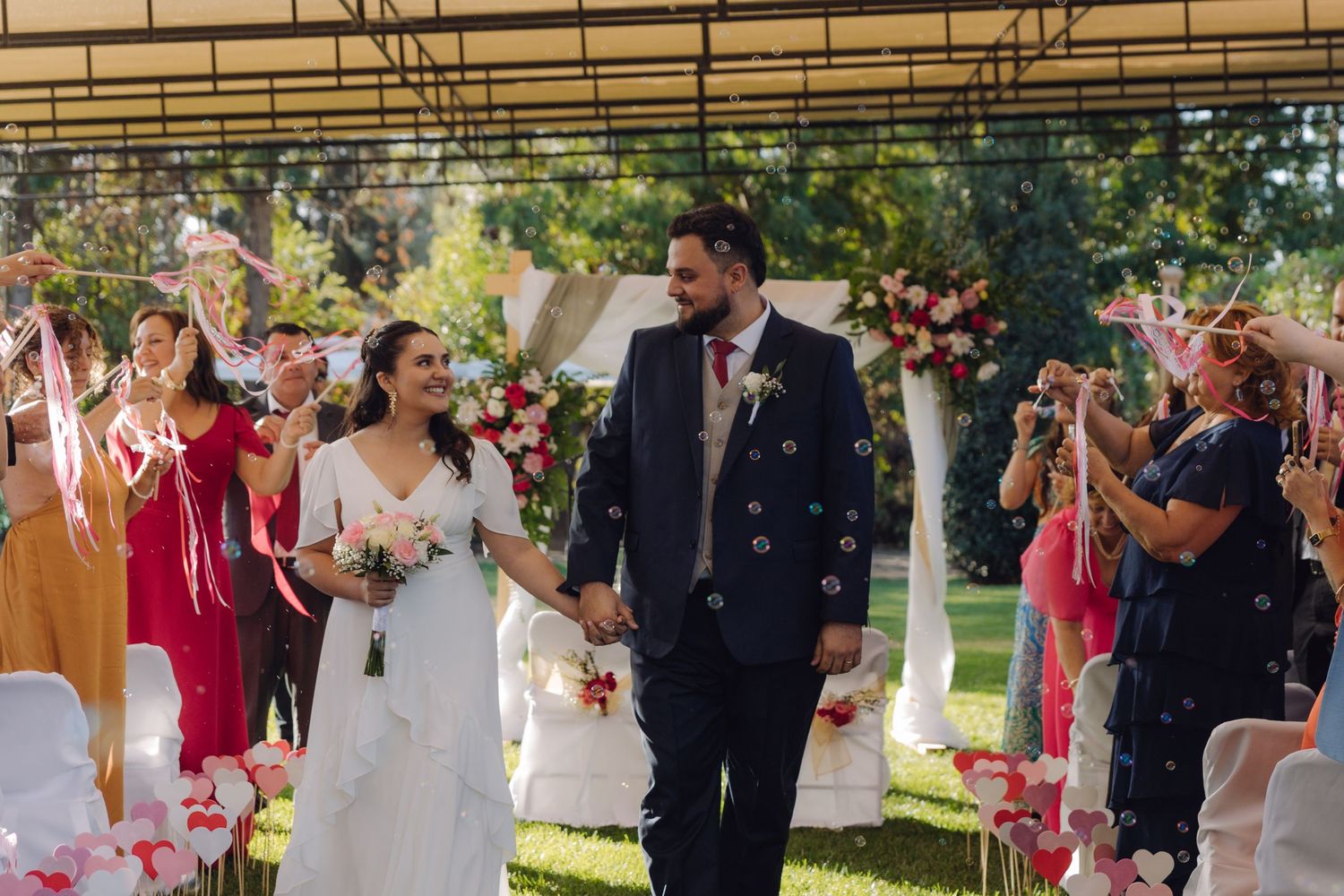 Bride and groom walk down outdoor wedding aisle as guests celebrate with confetti and streamers.