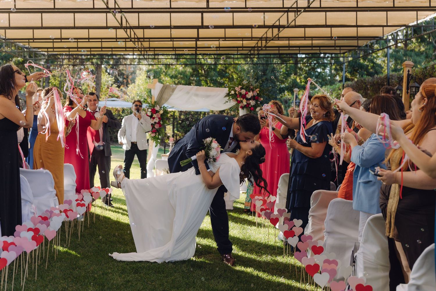 Groom dips bride in romantic kiss while walking down outdoor wedding aisle lined with cheering guests.