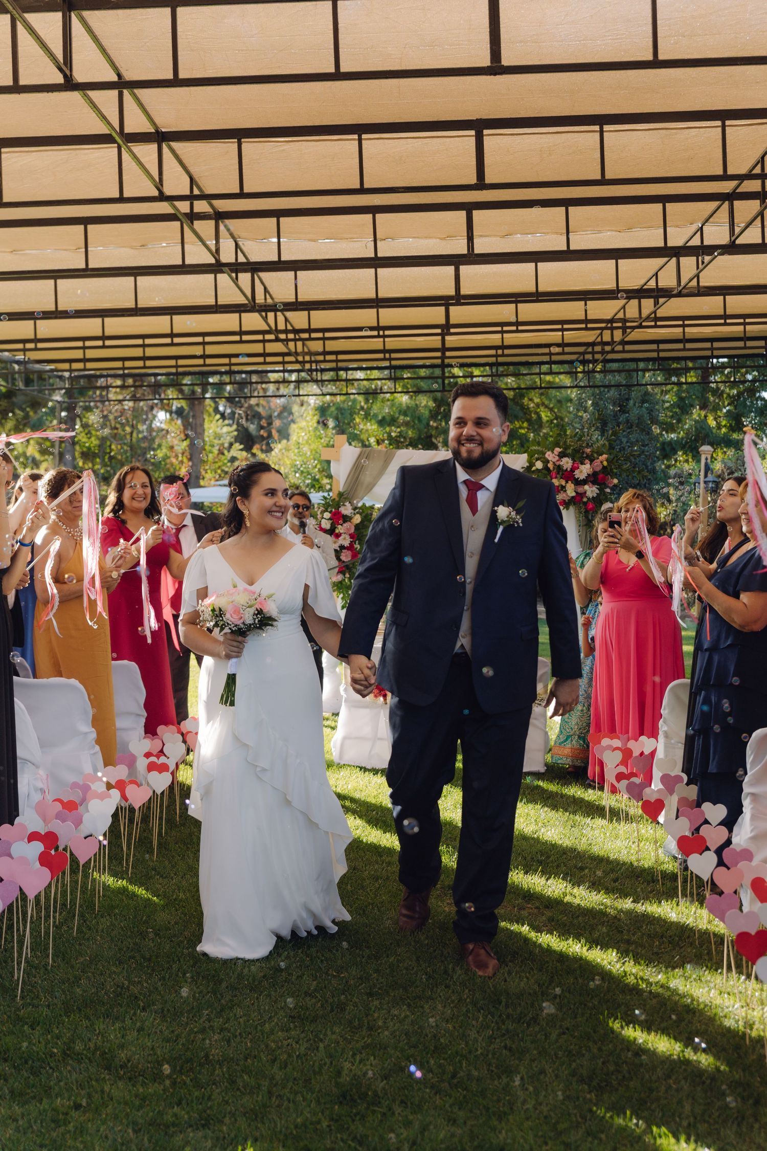 Smiling newlyweds hold hands walking down decorated outdoor wedding aisle with heart-shaped decorations.