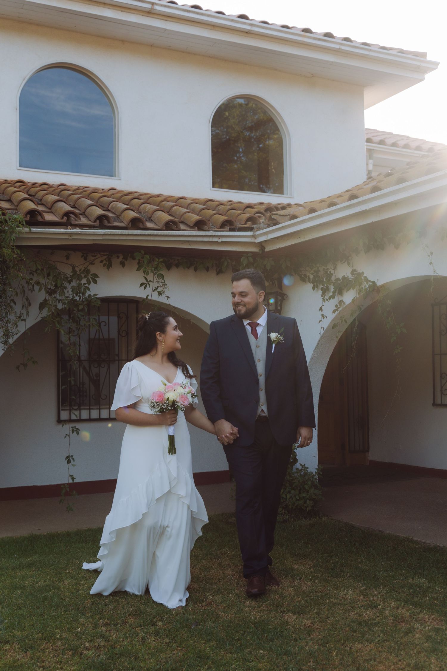 Bride and groom holding hands and smiling outside a Spanish-style white villa.