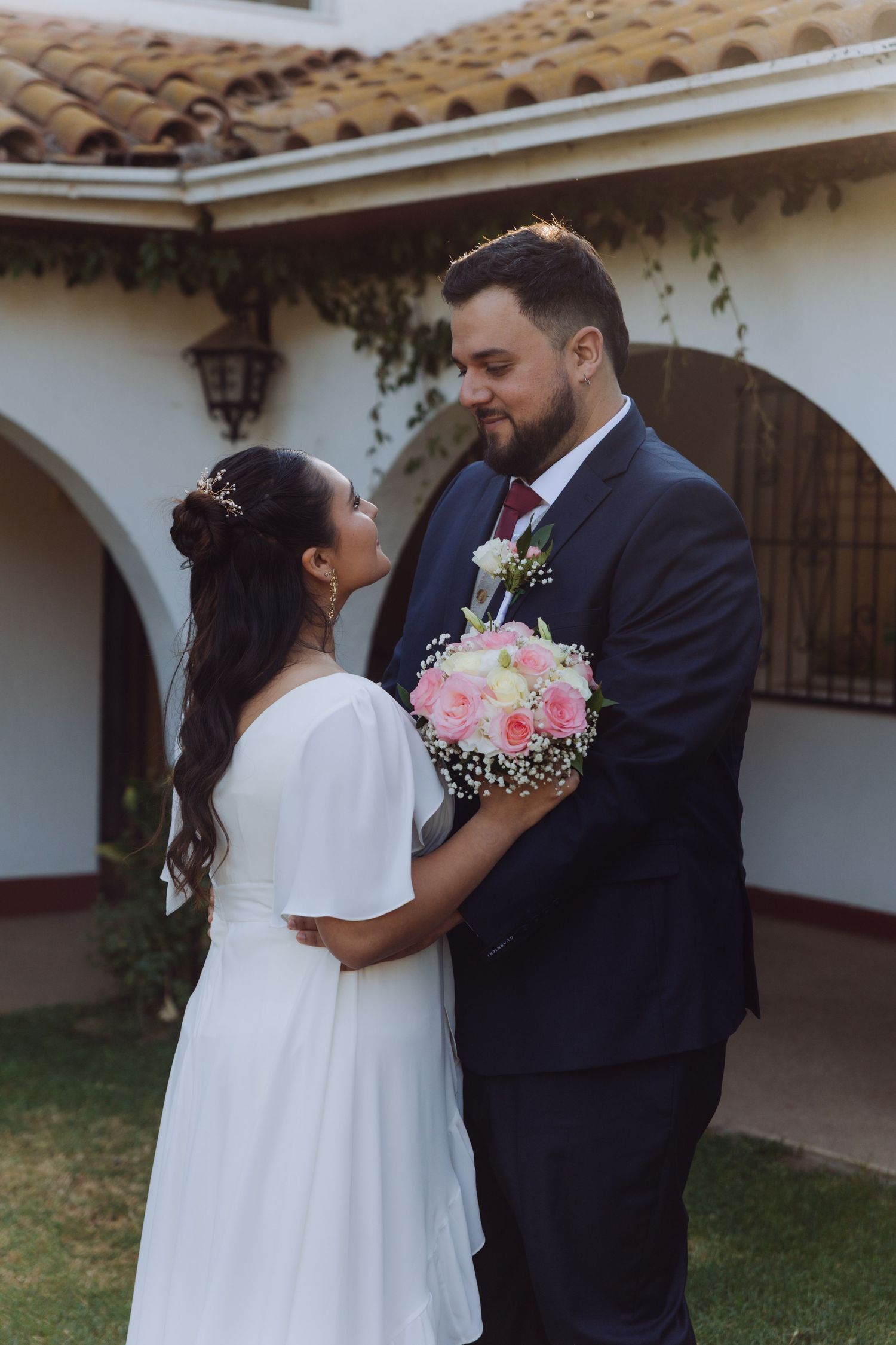 Groom gazing at bride holding pink rose bouquet in front of arched building.