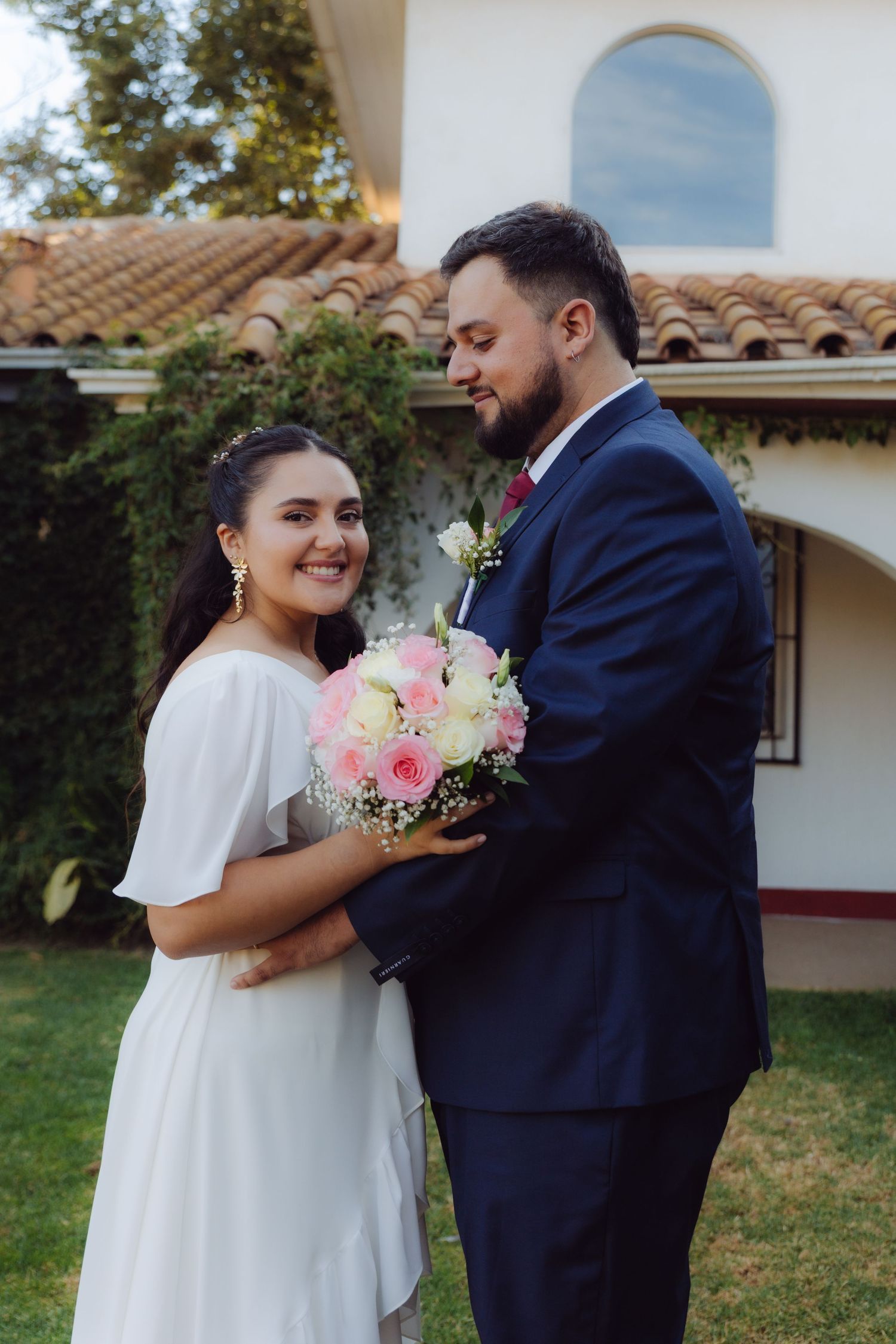 Smiling bride and groom posing together with pink bouquet outside Spanish villa.