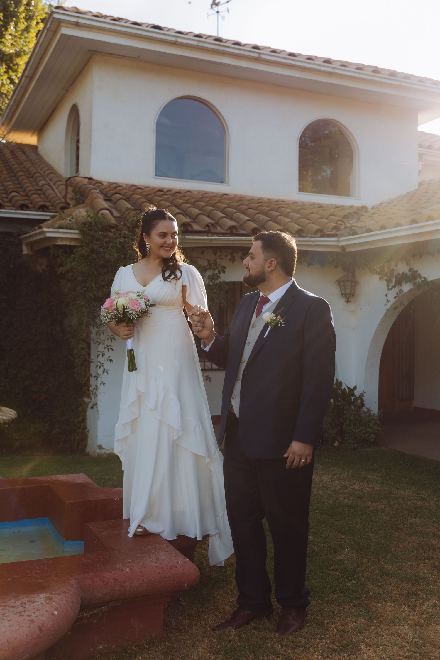 Bride in white gown holds bouquet while groom in dark suit holds her hand outside Spanish-style villa.