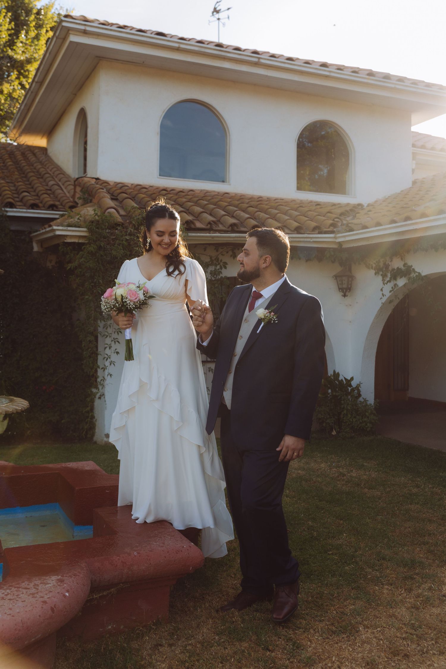 Wedding couple holding hands outside a Spanish-style white stucco home with terracotta roof tiles.