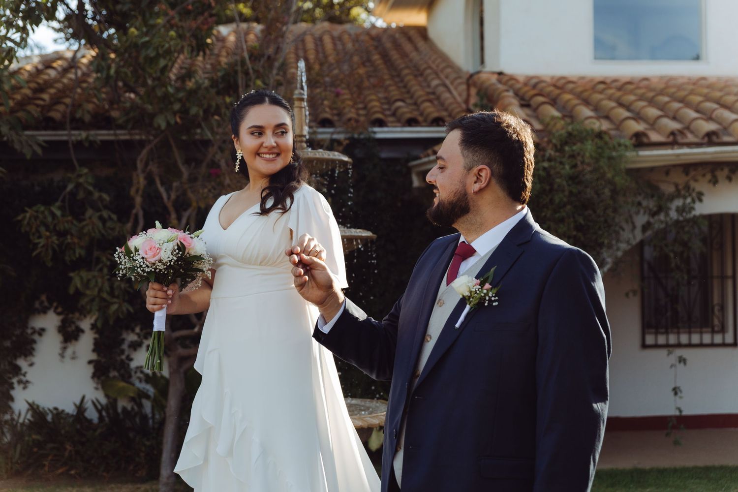 Smiling bride holding pink bouquet stands with groom in navy suit outside rustic venue in sunlight.