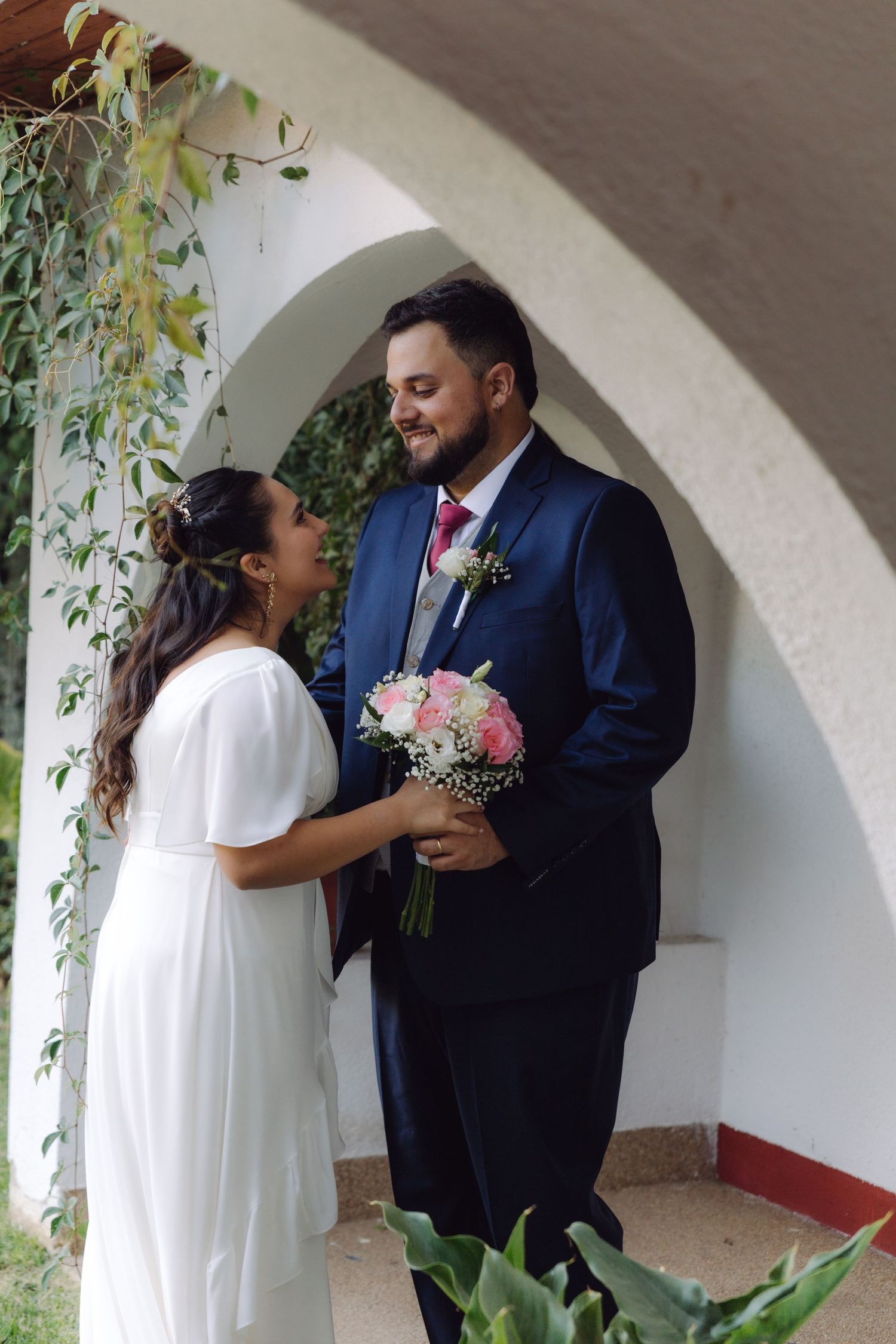 Bride and groom share intimate moment under white arched doorway with lush green vines overhead.