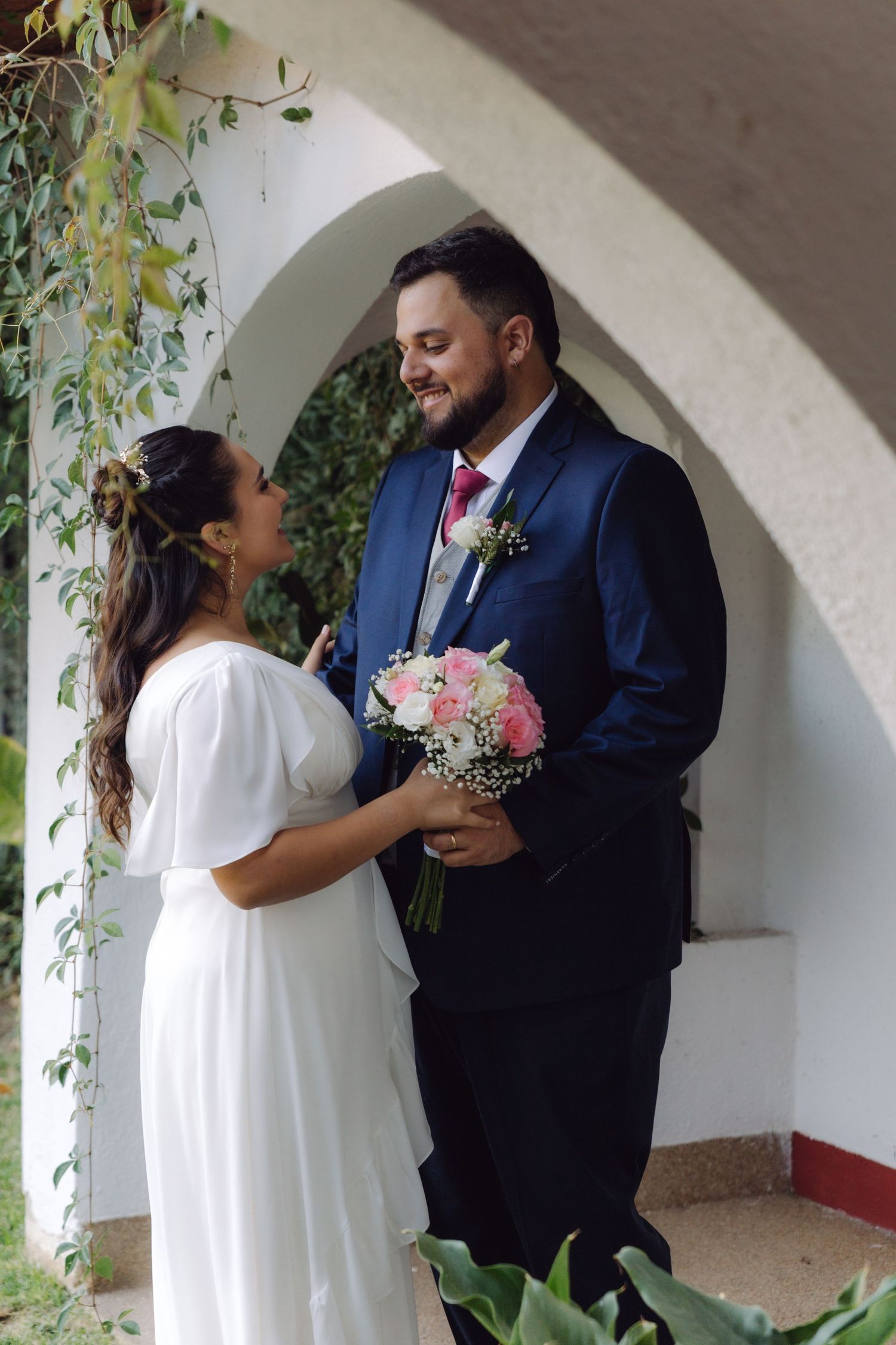 Newlyweds gazing at each other lovingly under a white arch surrounded by green foliage outdoors.