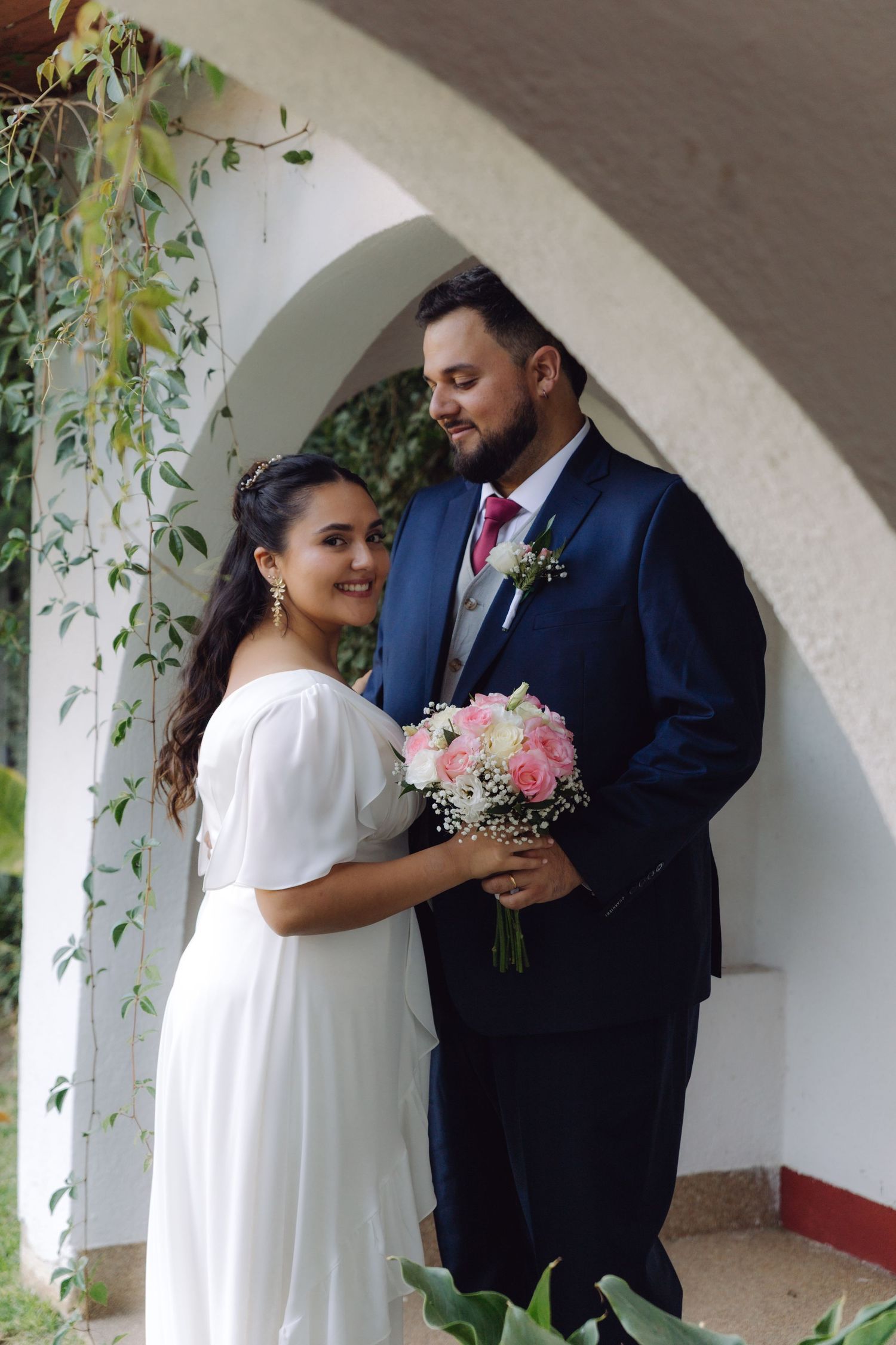 Bride in white dress holds pink bouquet while groom in navy suit smiles under elegant white arch.