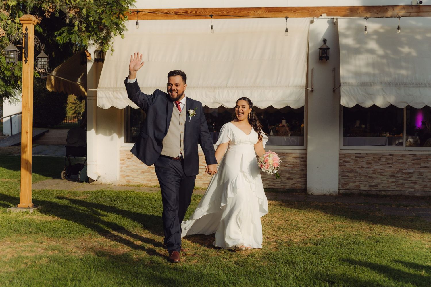 Joyful groom waves while walking hand-in-hand with bride across green lawn at outdoor wedding venue.