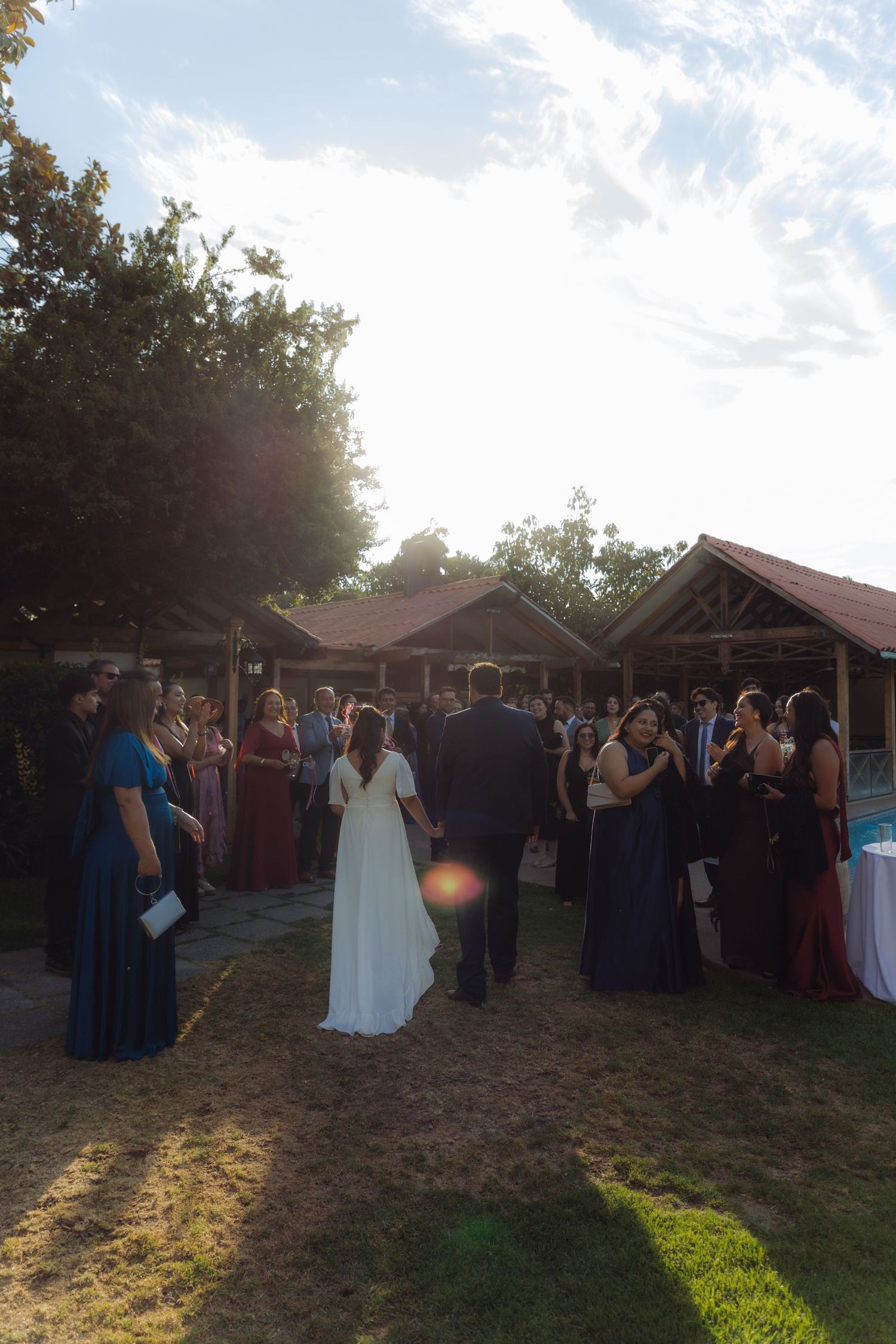 Wedding couple walks toward guests gathered under rustic wooden pavilion during outdoor garden ceremony.