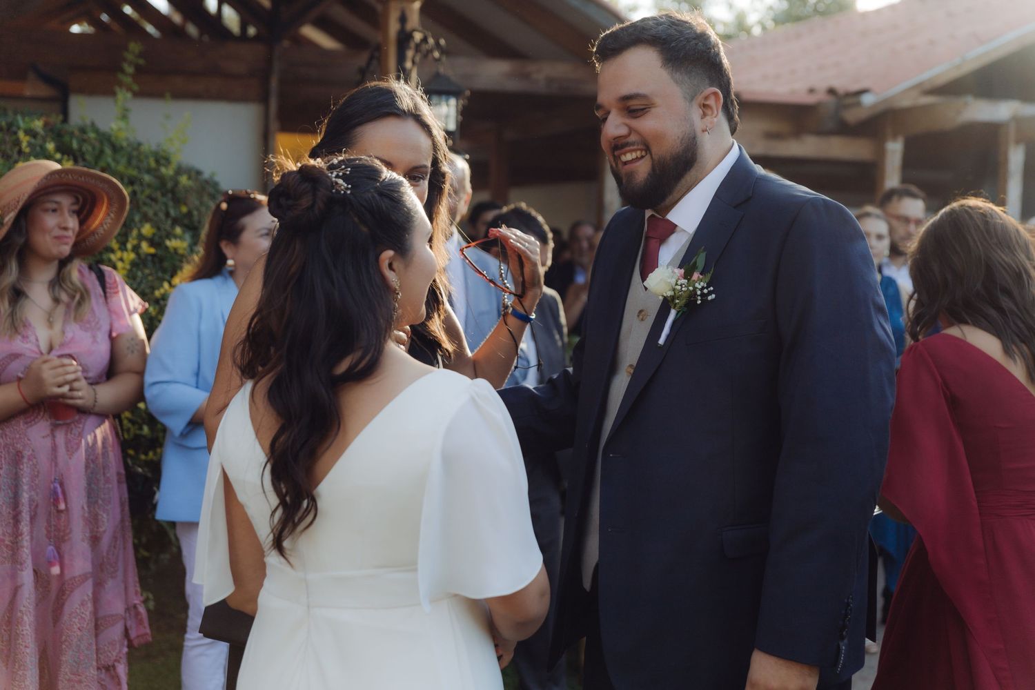 Groom in navy suit smiles warmly as bride faces him during outdoor wedding reception greeting guests.