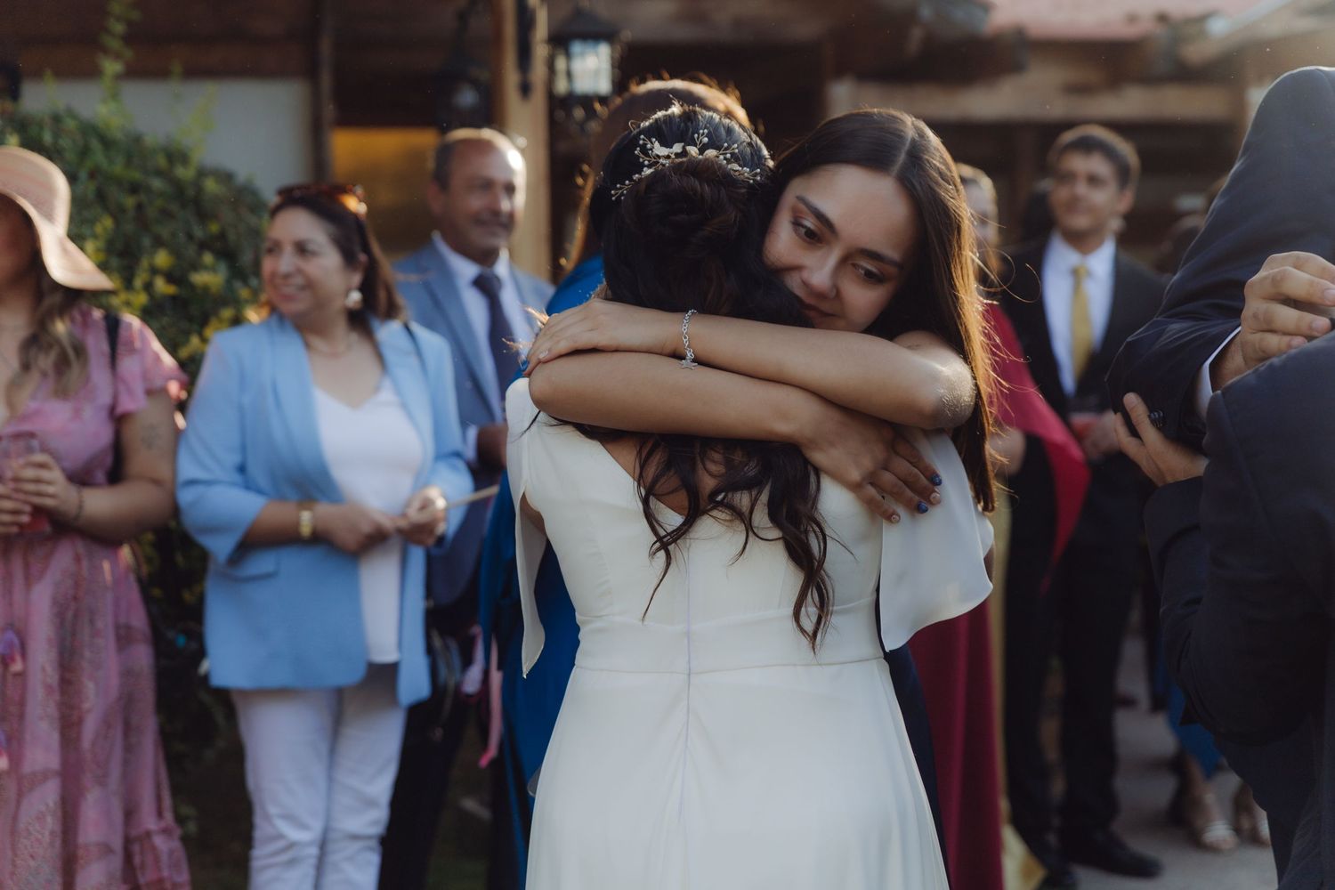 Bride in white gown embraces a guest warmly while wedding attendees celebrate in the background.