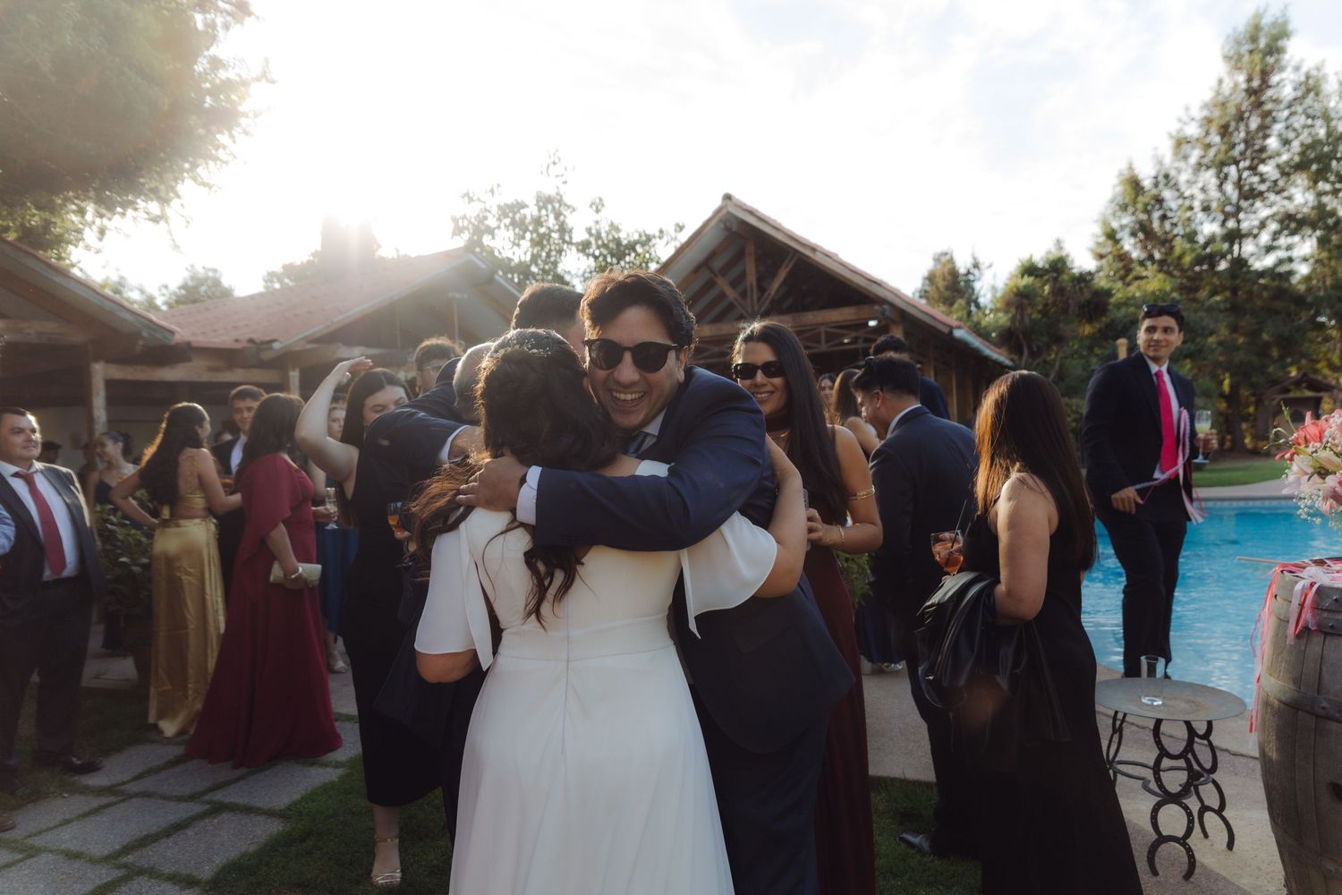 Bride in white dress hugging a guest at an outdoor wedding reception near a pool.