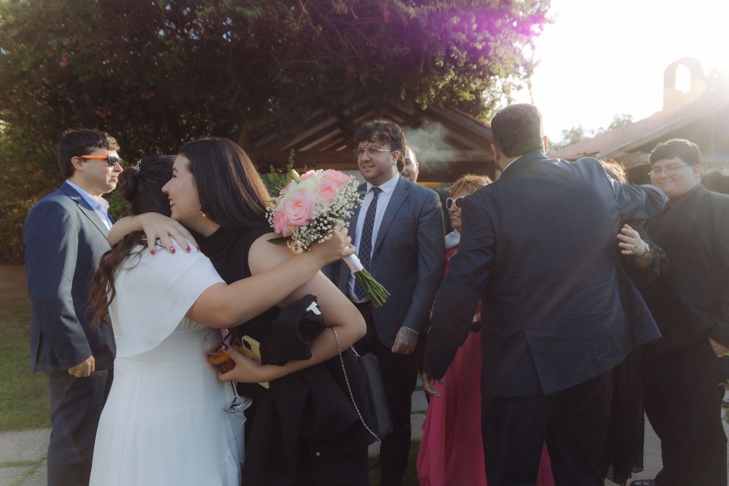 Bride holding pink bouquet embracing guests during a sunny outdoor wedding reception.