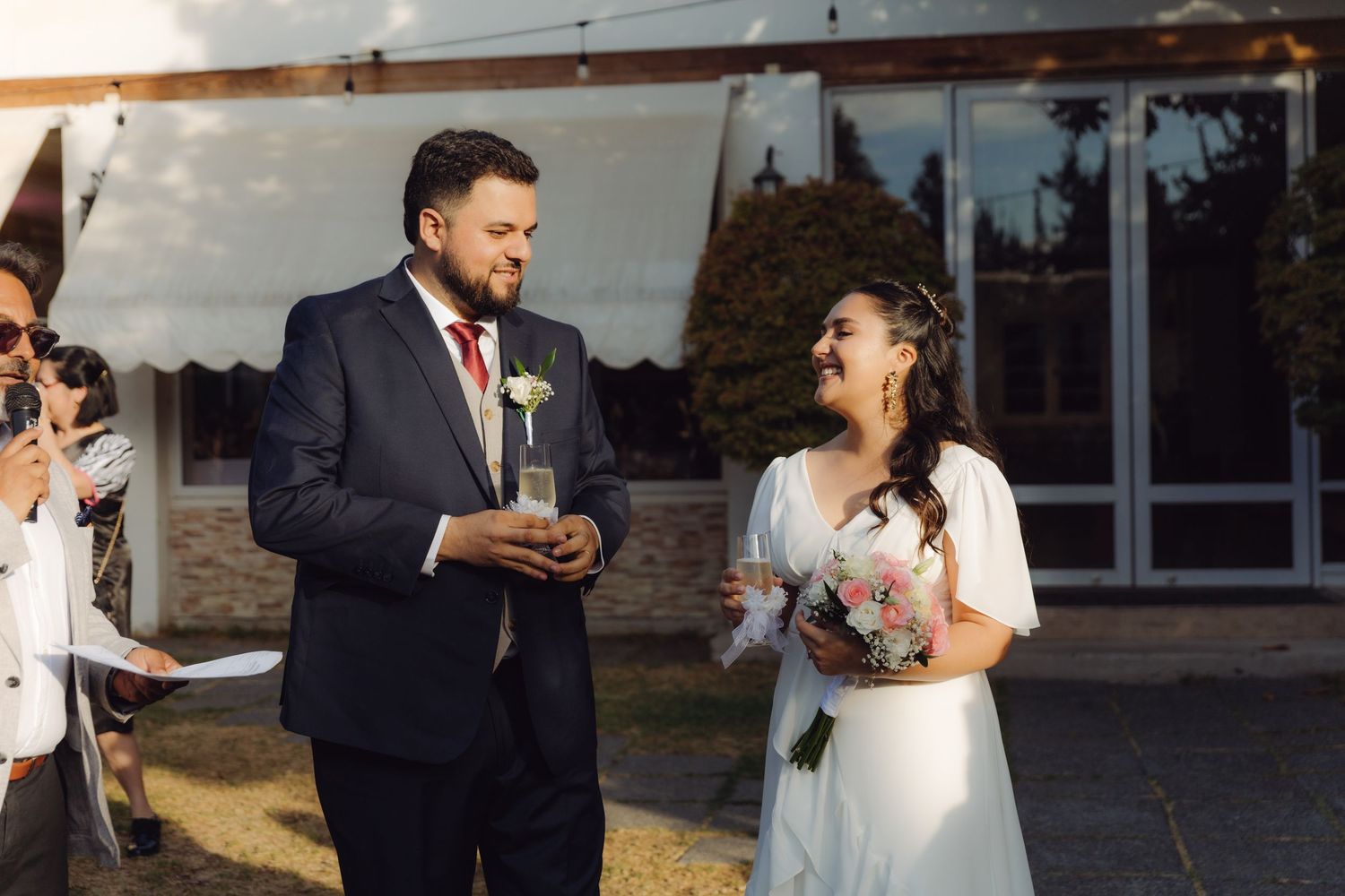 Smiling bride holding bouquet and groom exchanging vows at an outdoor wedding ceremony.