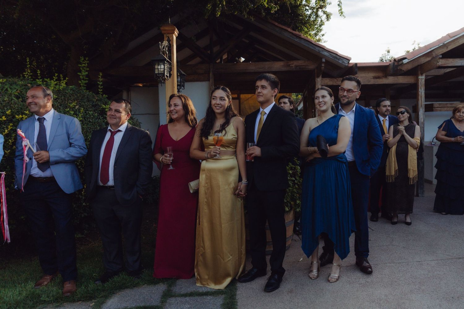 Wedding guests in formal attire standing in a row watching an outdoor ceremony.