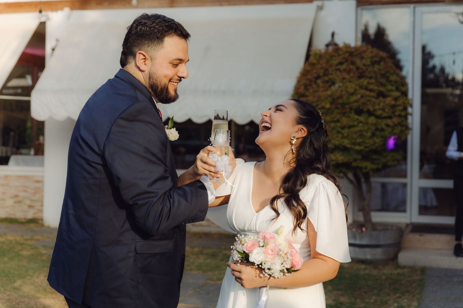 Joyful bride and groom clinking champagne glasses and laughing at their outdoor wedding.
