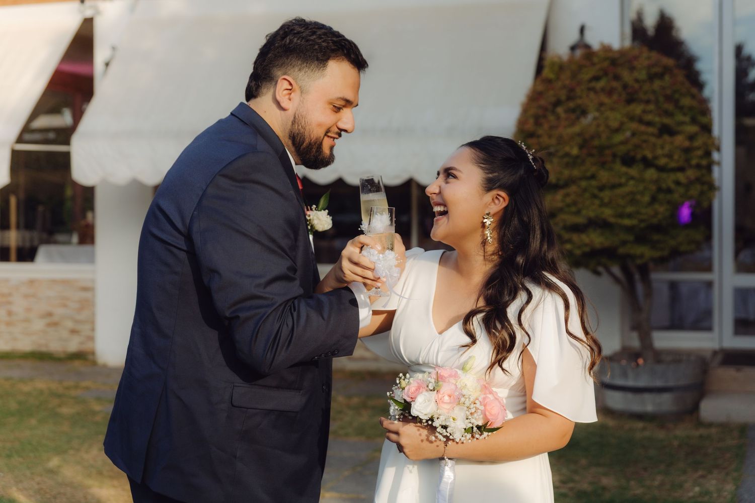 Joyful newlyweds toasting with champagne glasses outdoors, bride holding pink bouquet in white dress, groom in navy suit.