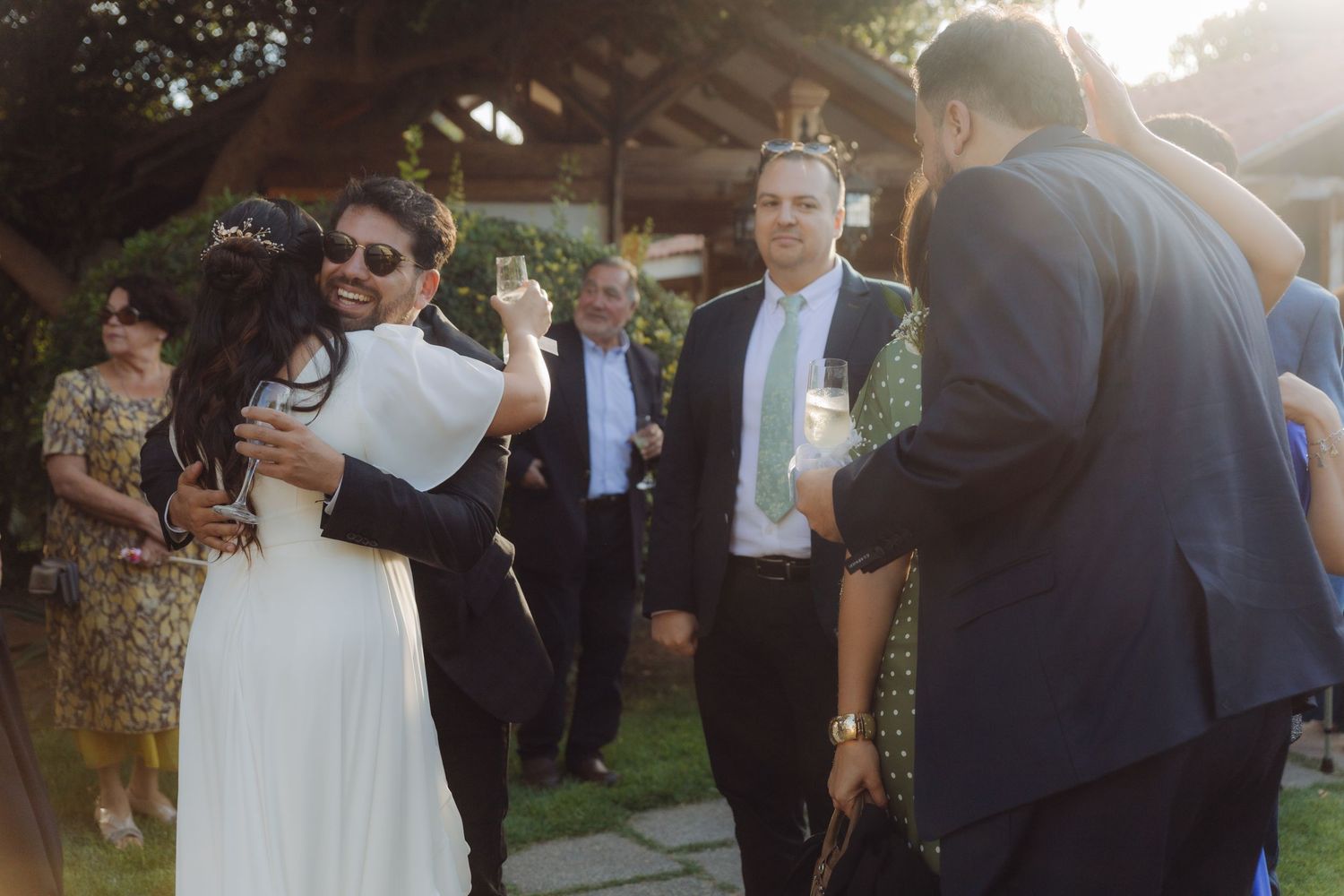 Wedding guests embracing and celebrating outdoors at golden hour, bride in white dress surrounded by well-wishers.