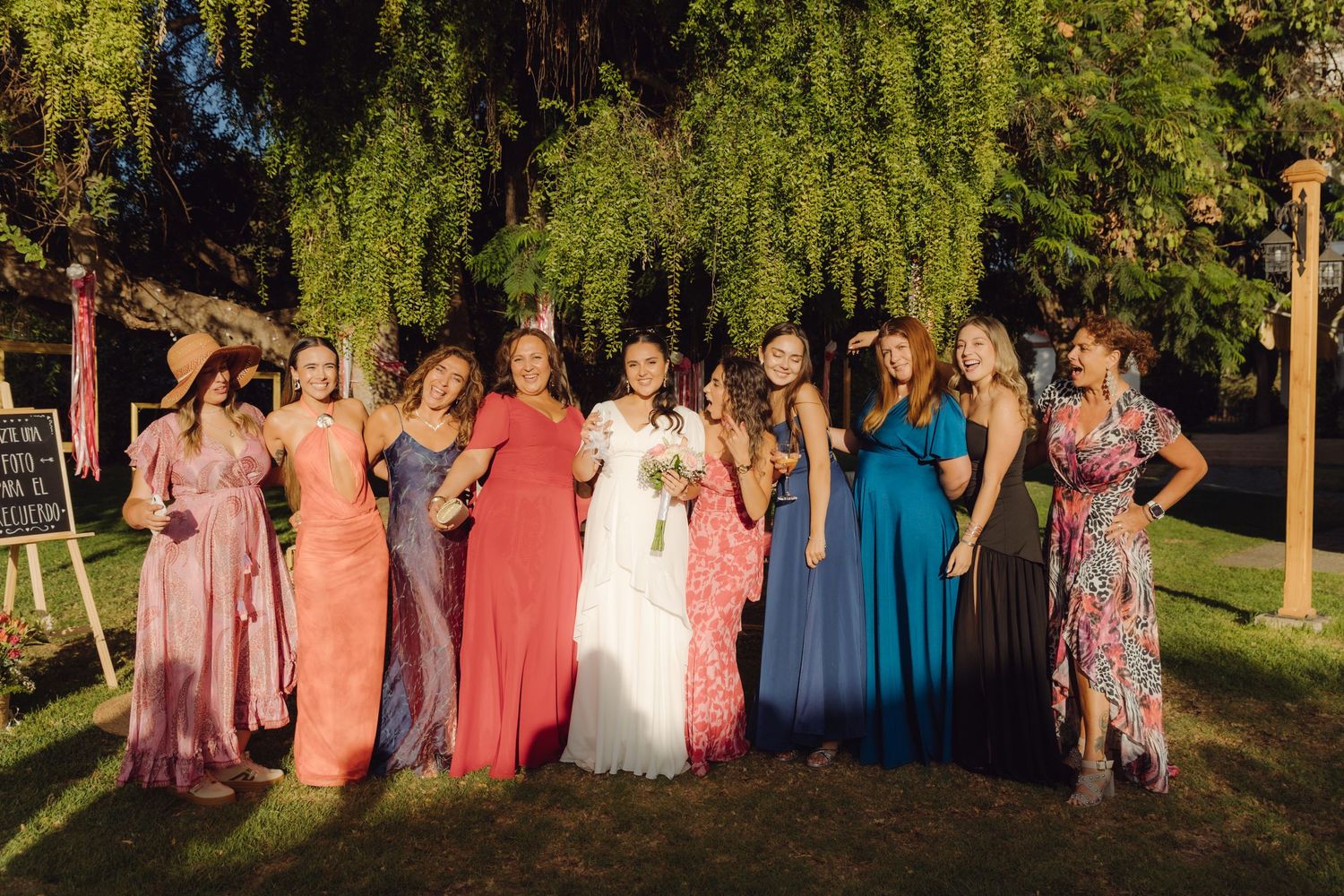 Smiling bride and bridesmaids in colorful formal gowns laughing together under lush green weeping willow tree.
