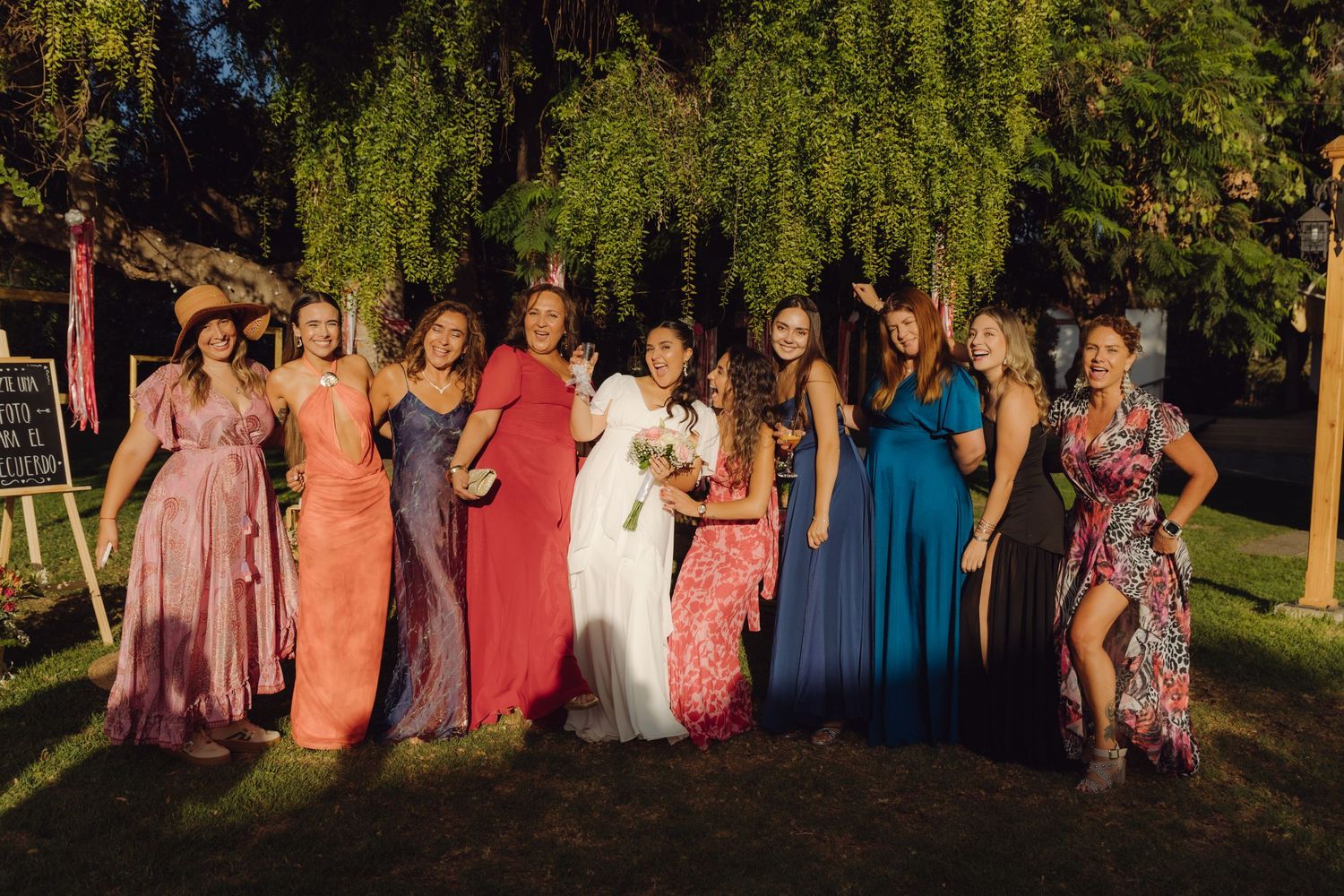 Bride posing with colorful bridesmaids in vibrant gowns under a weeping willow tree at outdoor wedding reception.