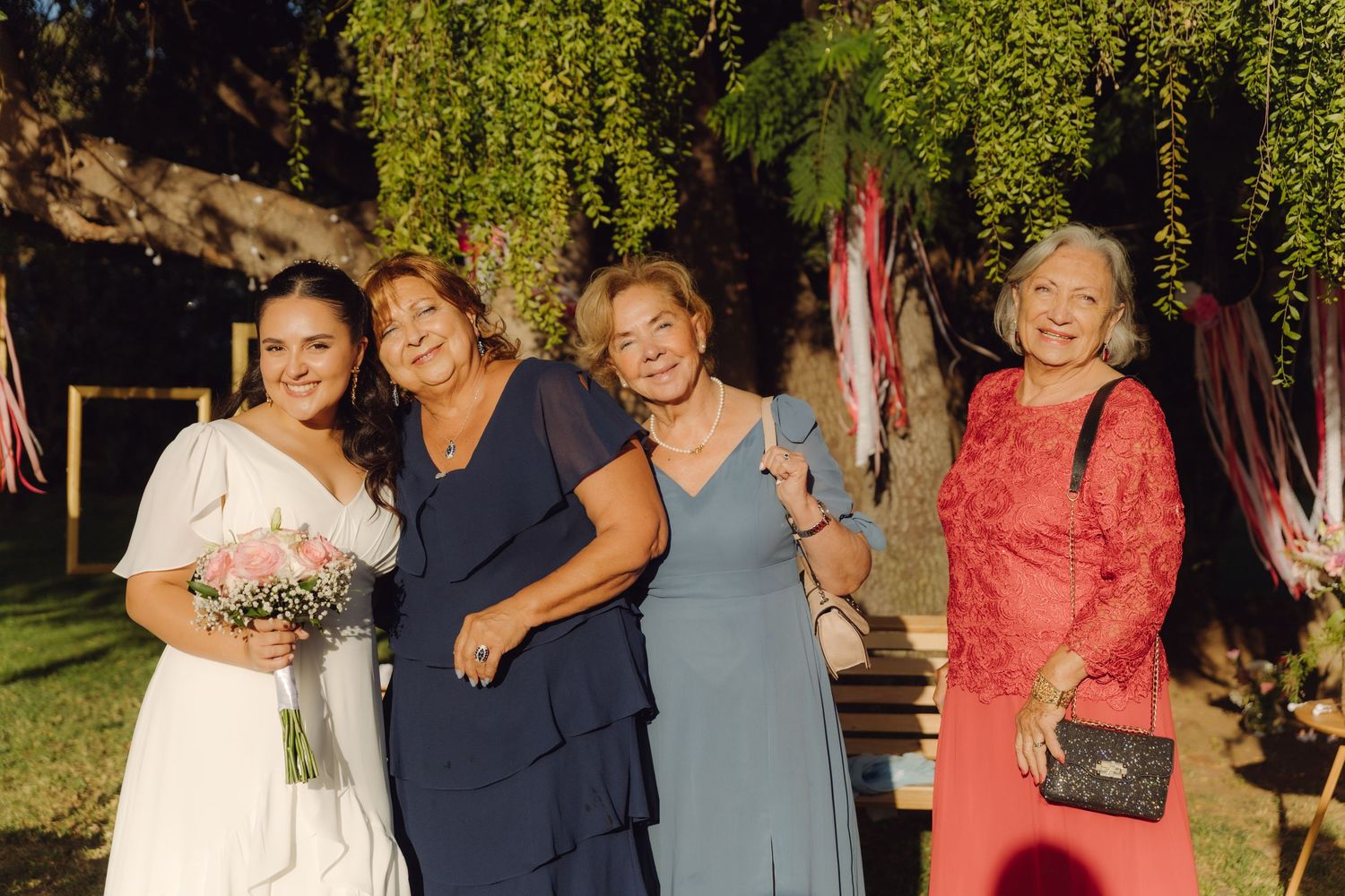 Bride with pink bouquet posing with three older female relatives in blue and red dresses at outdoor wedding.