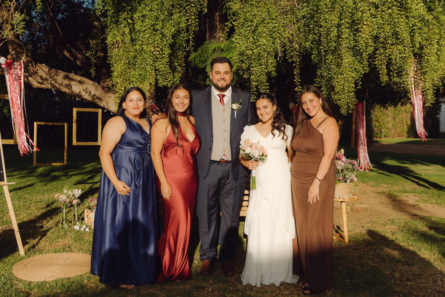 Wedding couple posing with four female guests in elegant colored gowns under a weeping willow tree outdoors.