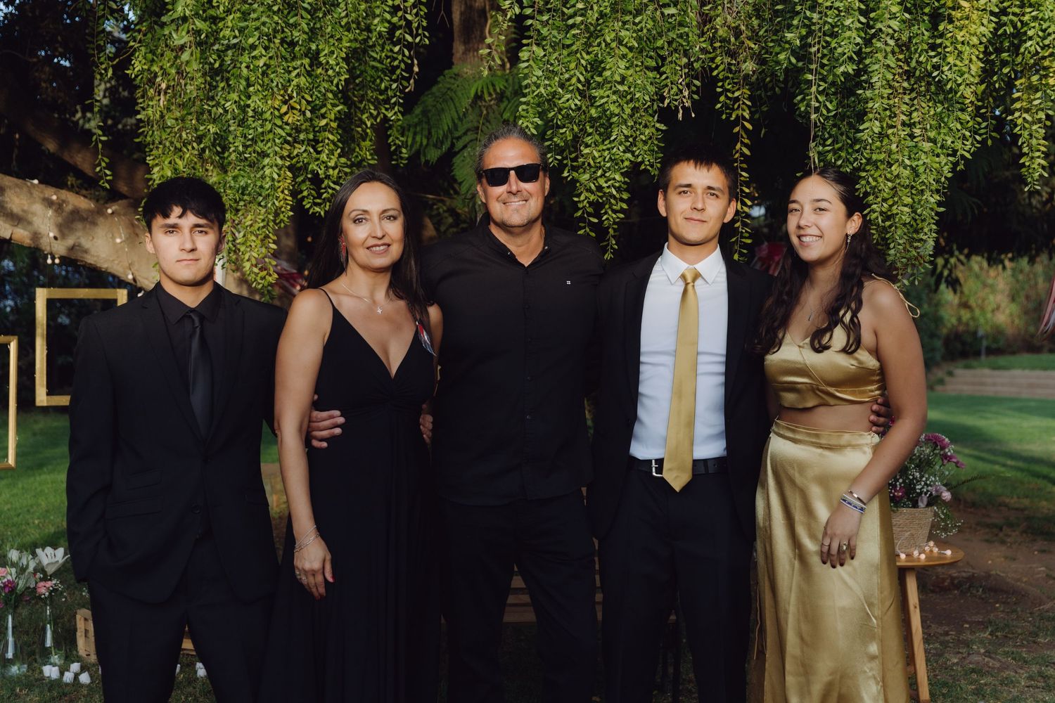 Family of five dressed in formal black and gold attire posing together at outdoor wedding reception at dusk.
