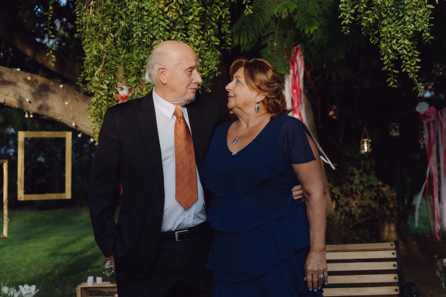 Elegant older couple sharing a tender glance at outdoor wedding reception surrounded by garden greenery at night.