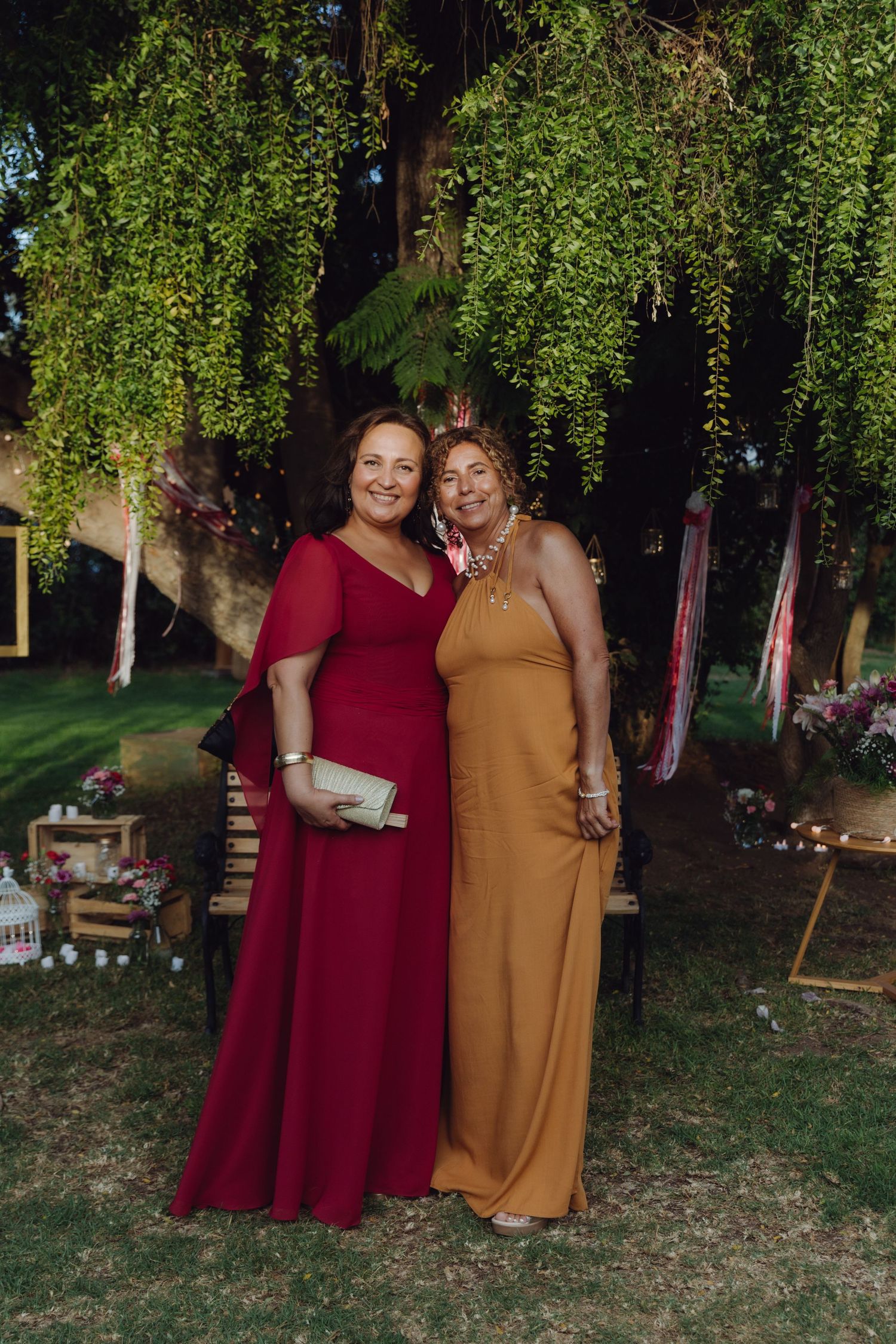 Two smiling women in elegant red and mustard formal gowns posing under a lush weeping willow at wedding.