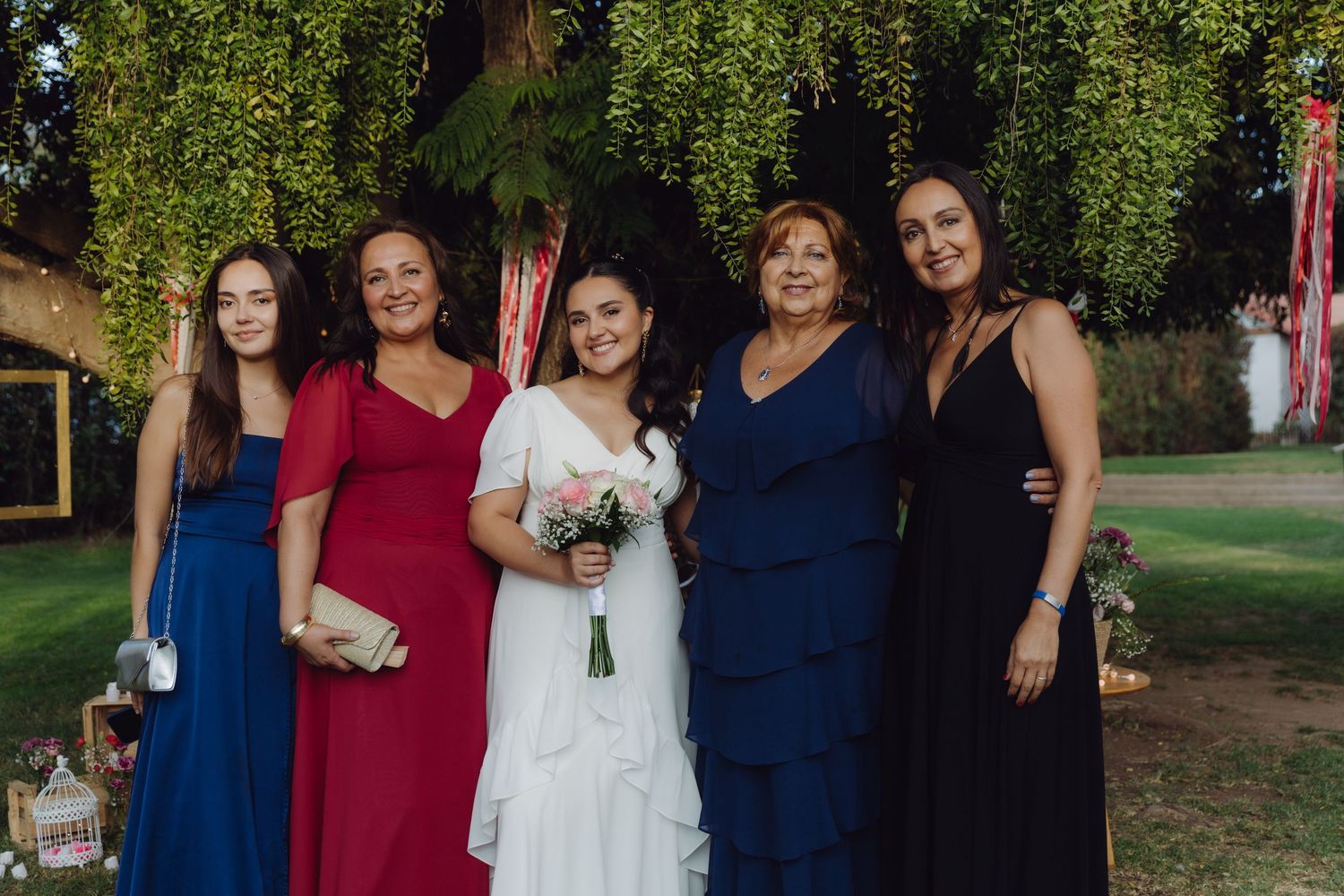 Bride holding bouquet posing with four female family members in blue, red, and black dresses at outdoor wedding.
