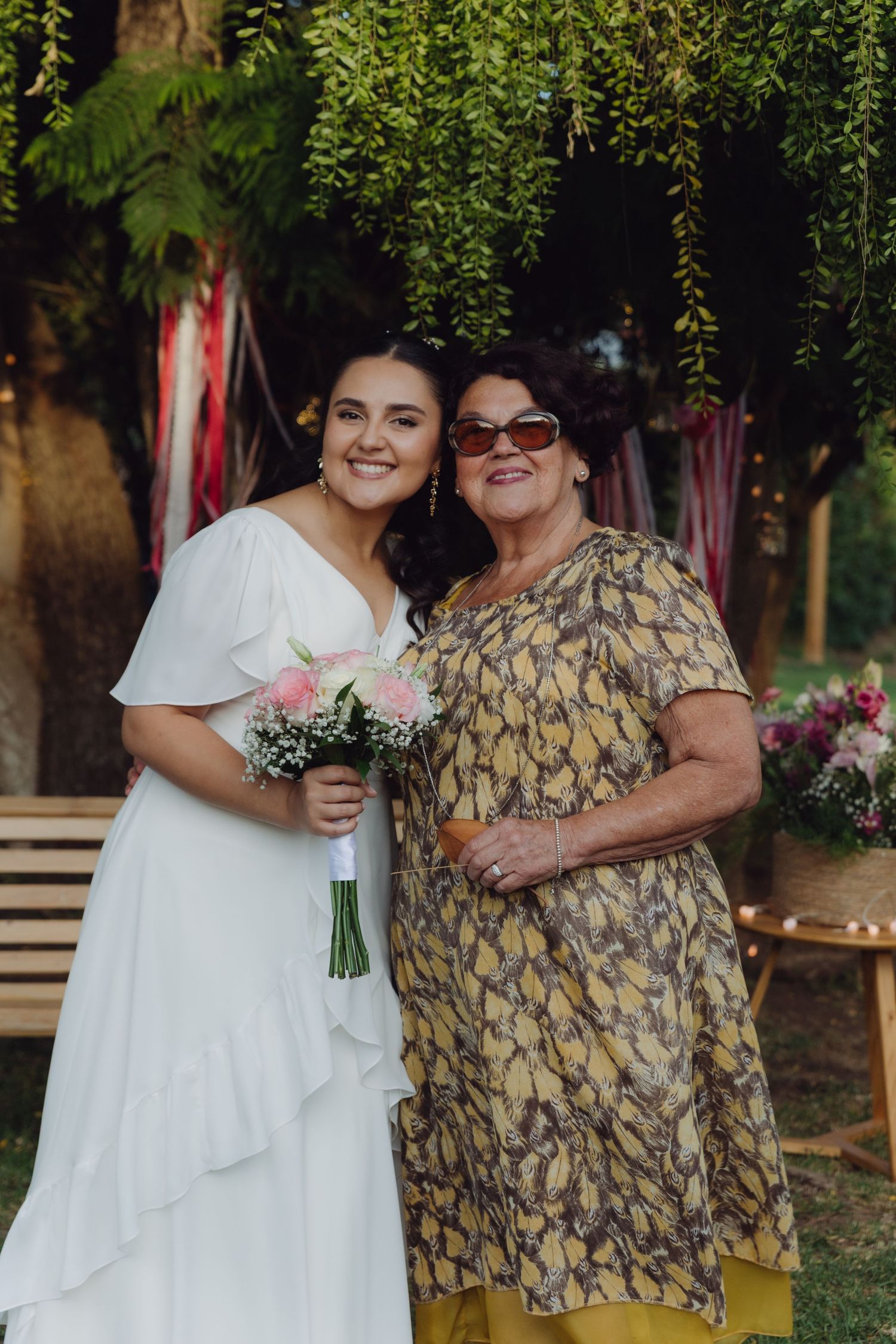 Bride in white dress holding pink bouquet poses with older woman in floral dress at outdoor wedding.