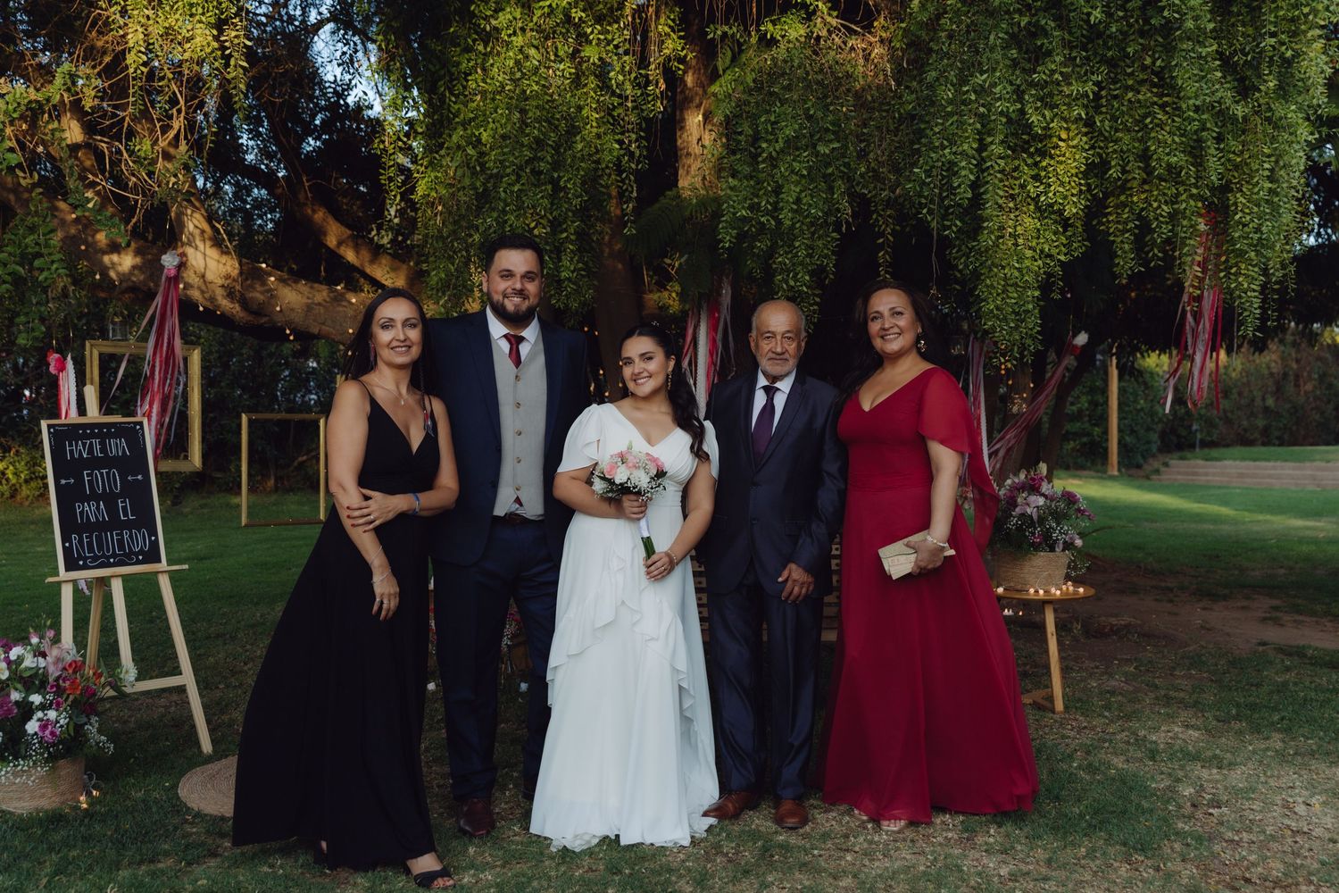 Wedding party of five poses outdoors under weeping tree with colorful decorations and chalkboard sign.