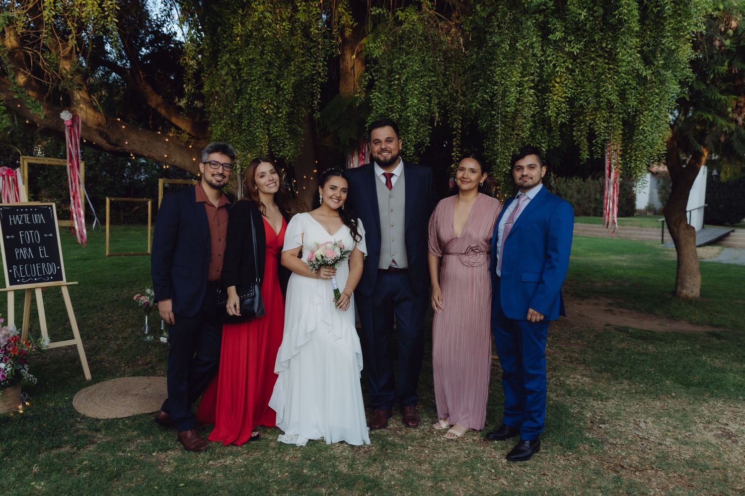 Six wedding guests in formal attire pose together outdoors under a tree at a garden wedding.