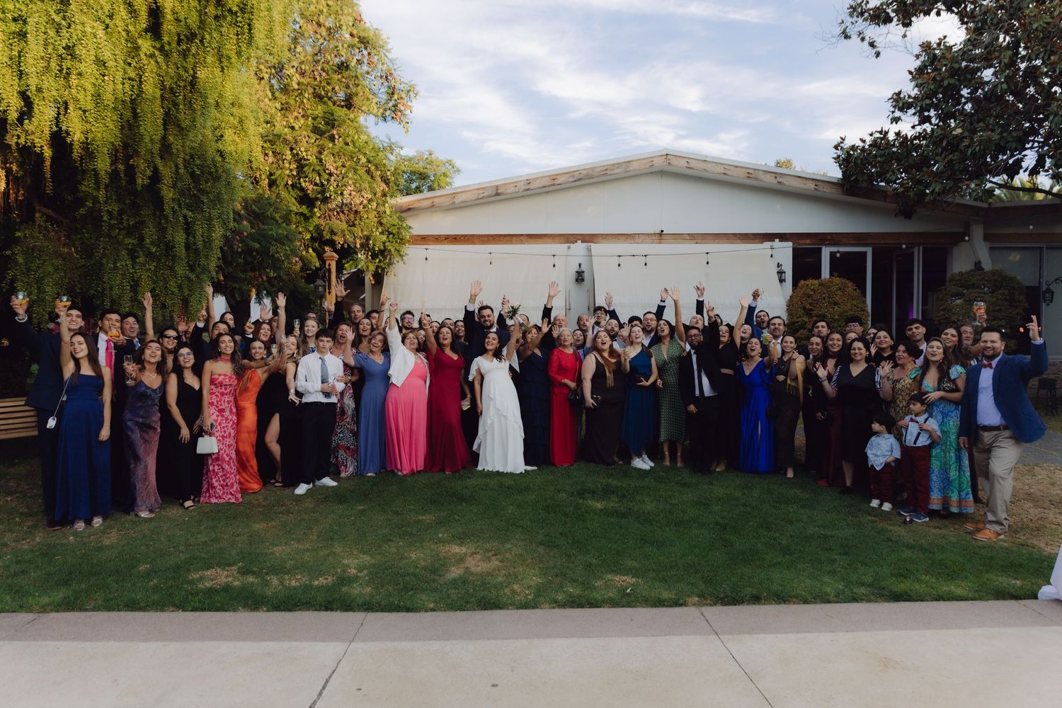 Large wedding celebration group photo with guests in colorful formal wear on a green lawn.