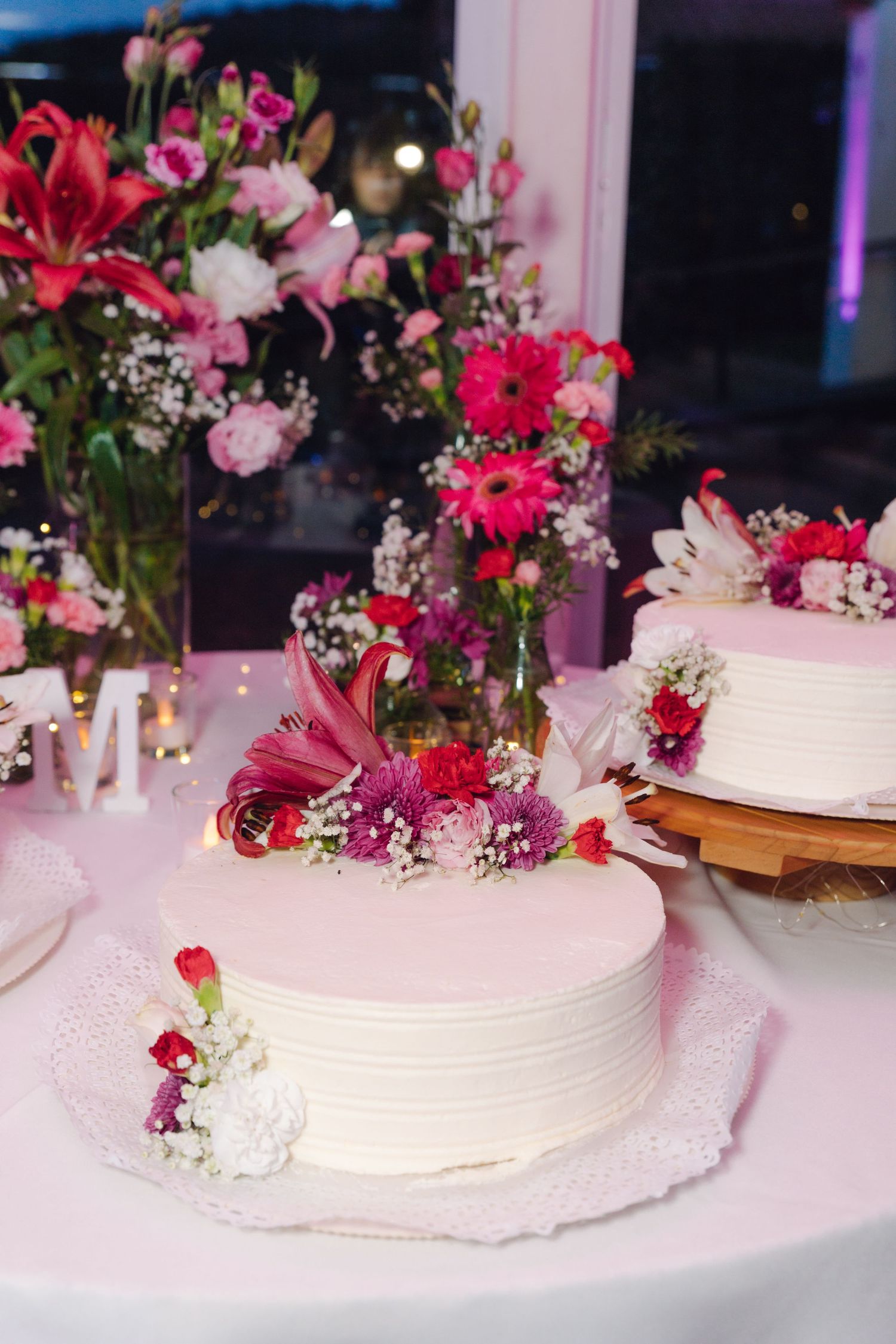 Elegant white wedding cake adorned with pink and red flowers beside floral arrangements.