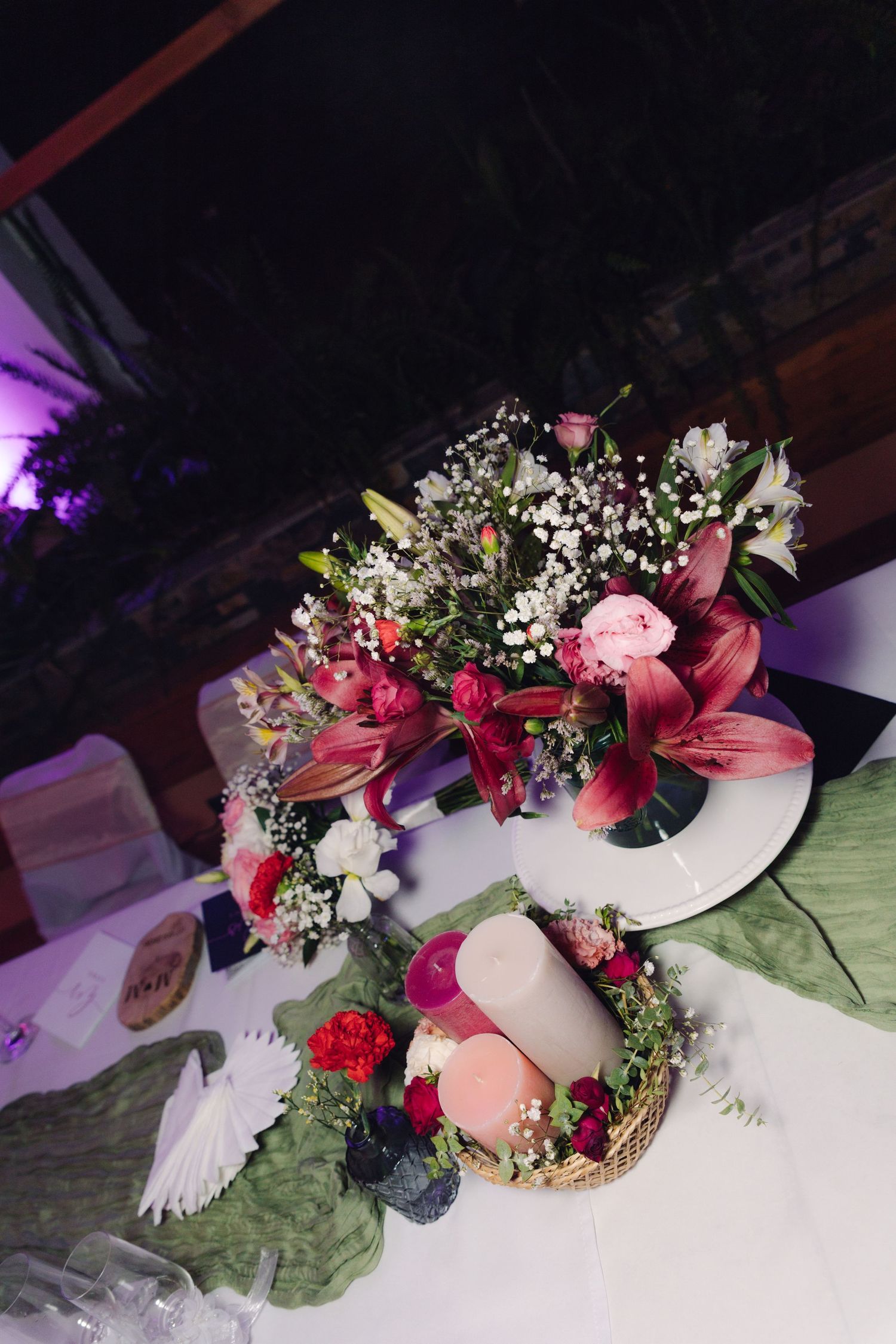 Wedding table centerpiece featuring pink lilies, roses, and candles on a white tablecloth.