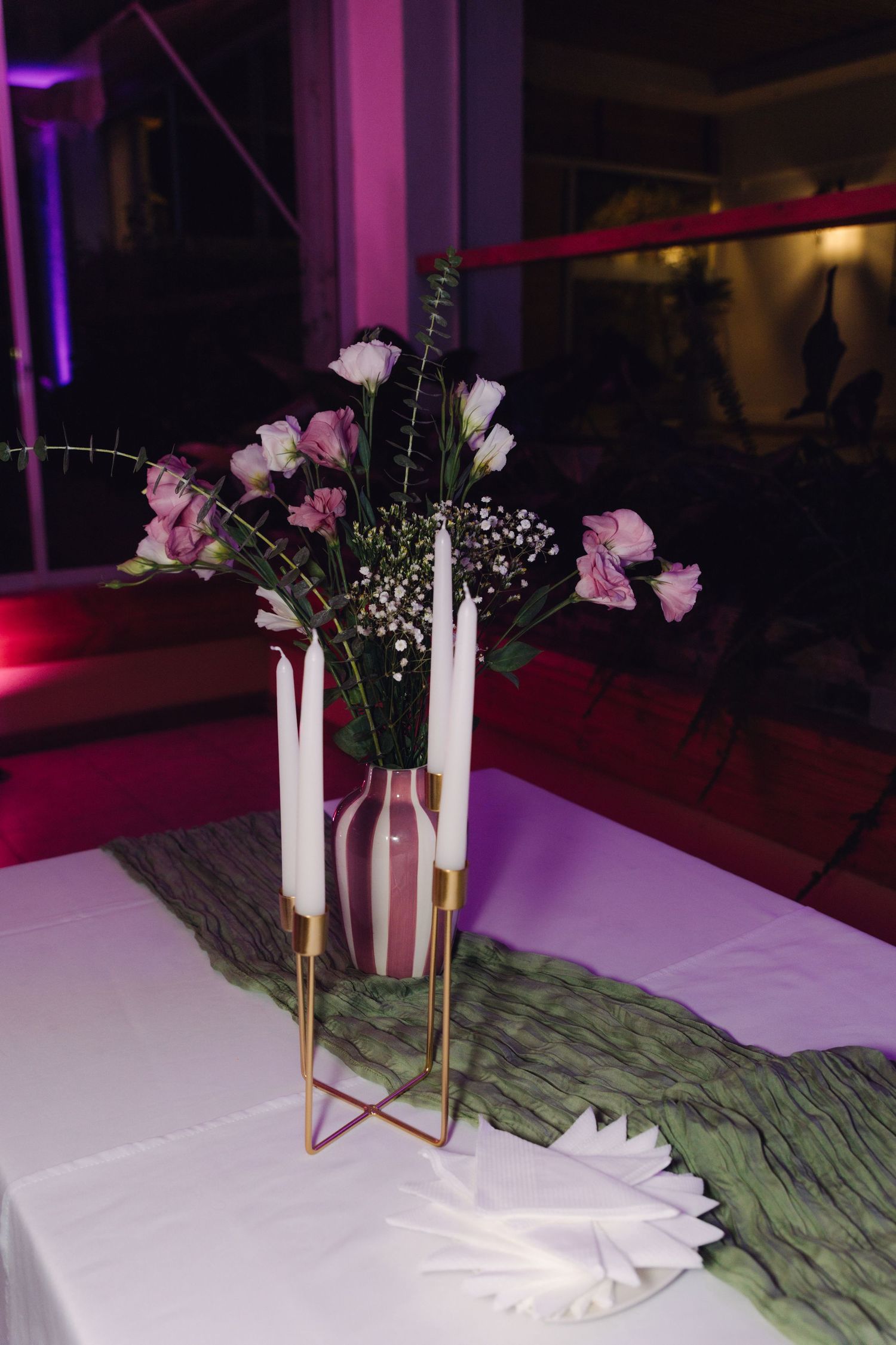 Wedding table centerpiece with white candles, striped vase, and pink flowers on green runner.