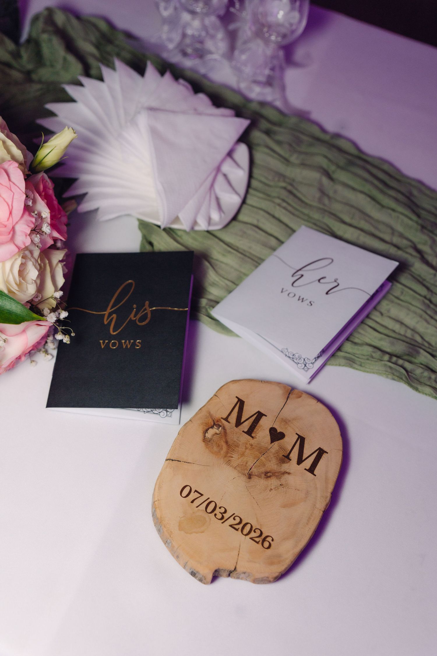 Wedding vow books labeled his and vows beside wooden slice engraved with M&M and 07/03/2026.