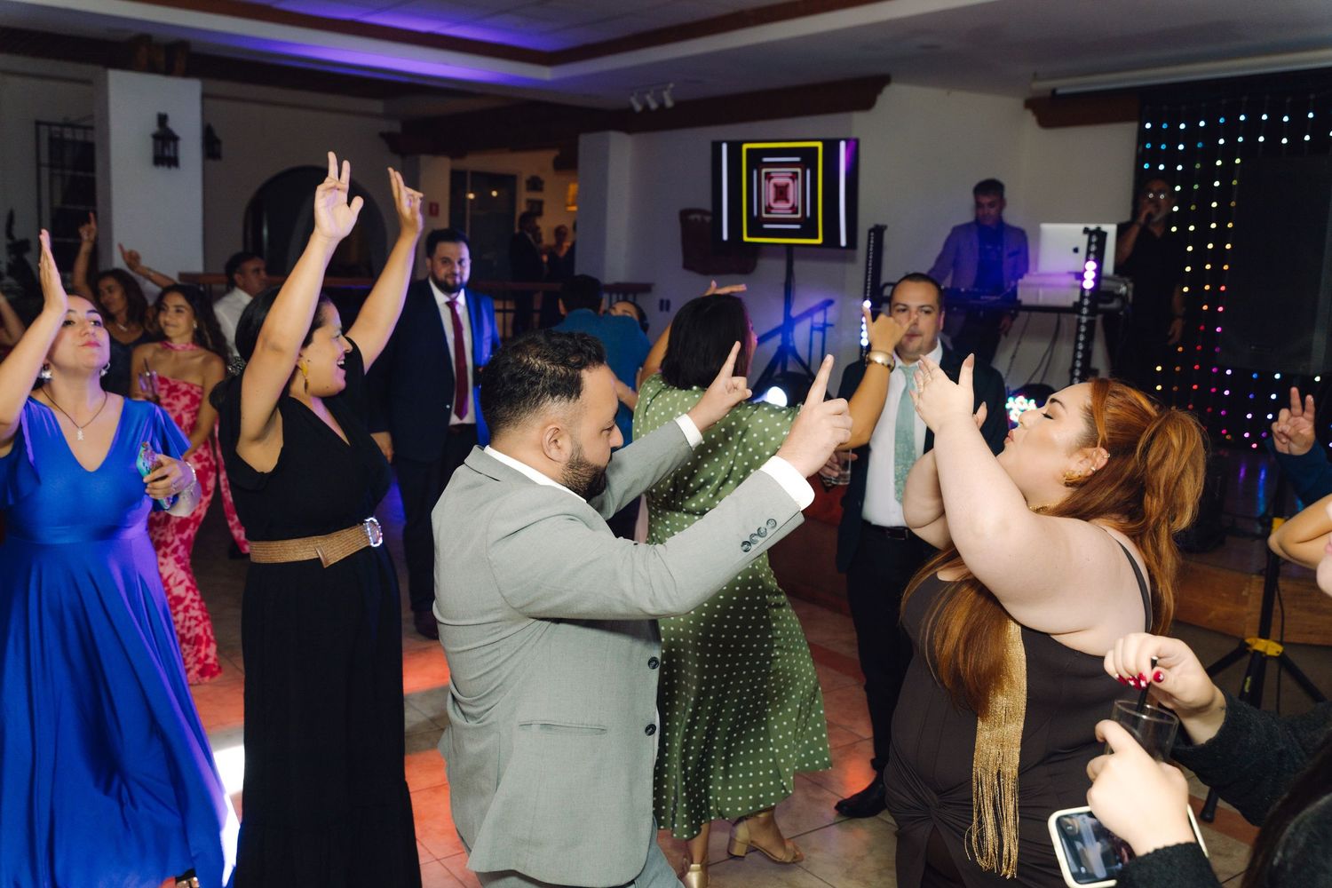 Excited wedding guests dancing and raising hands at a lively indoor wedding reception party.