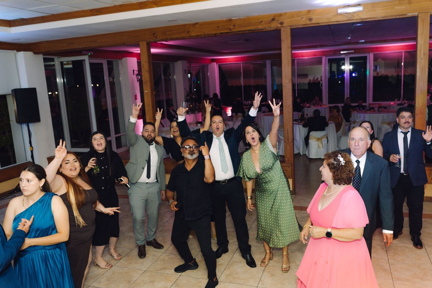 Large group of joyful wedding guests cheering with raised hands on the dance floor.