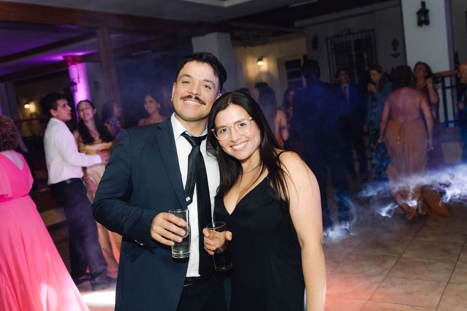 A couple poses together at a wedding reception, man with mustache in suit holding drink.