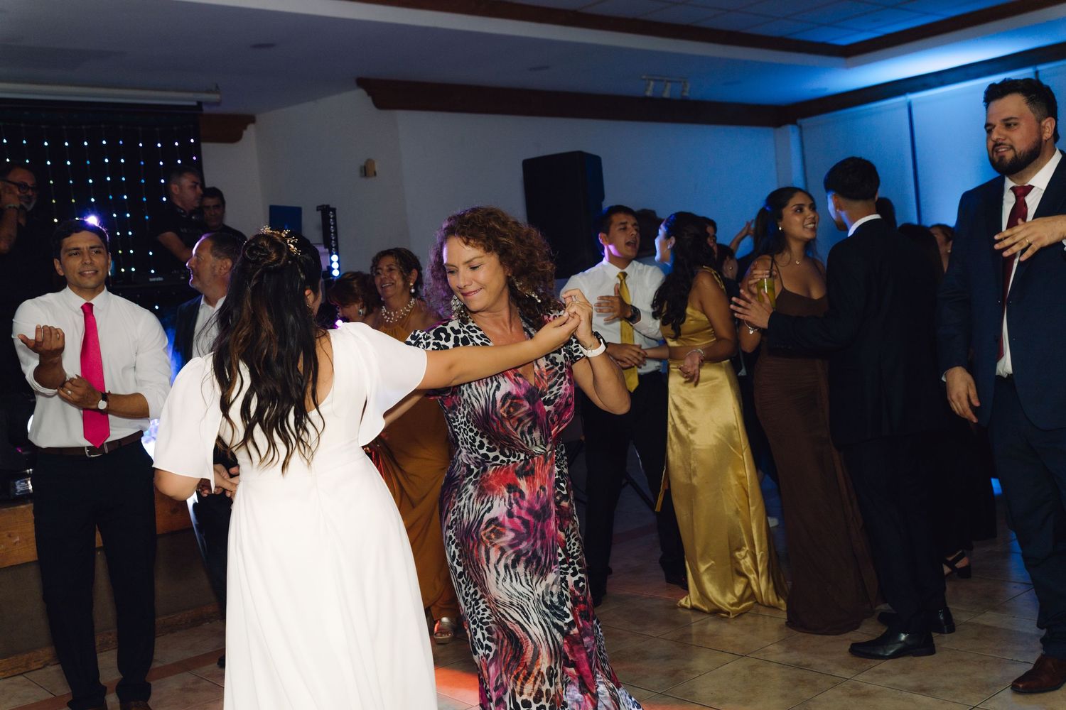Bride in white dress holds hands dancing with a woman in floral dress at reception.