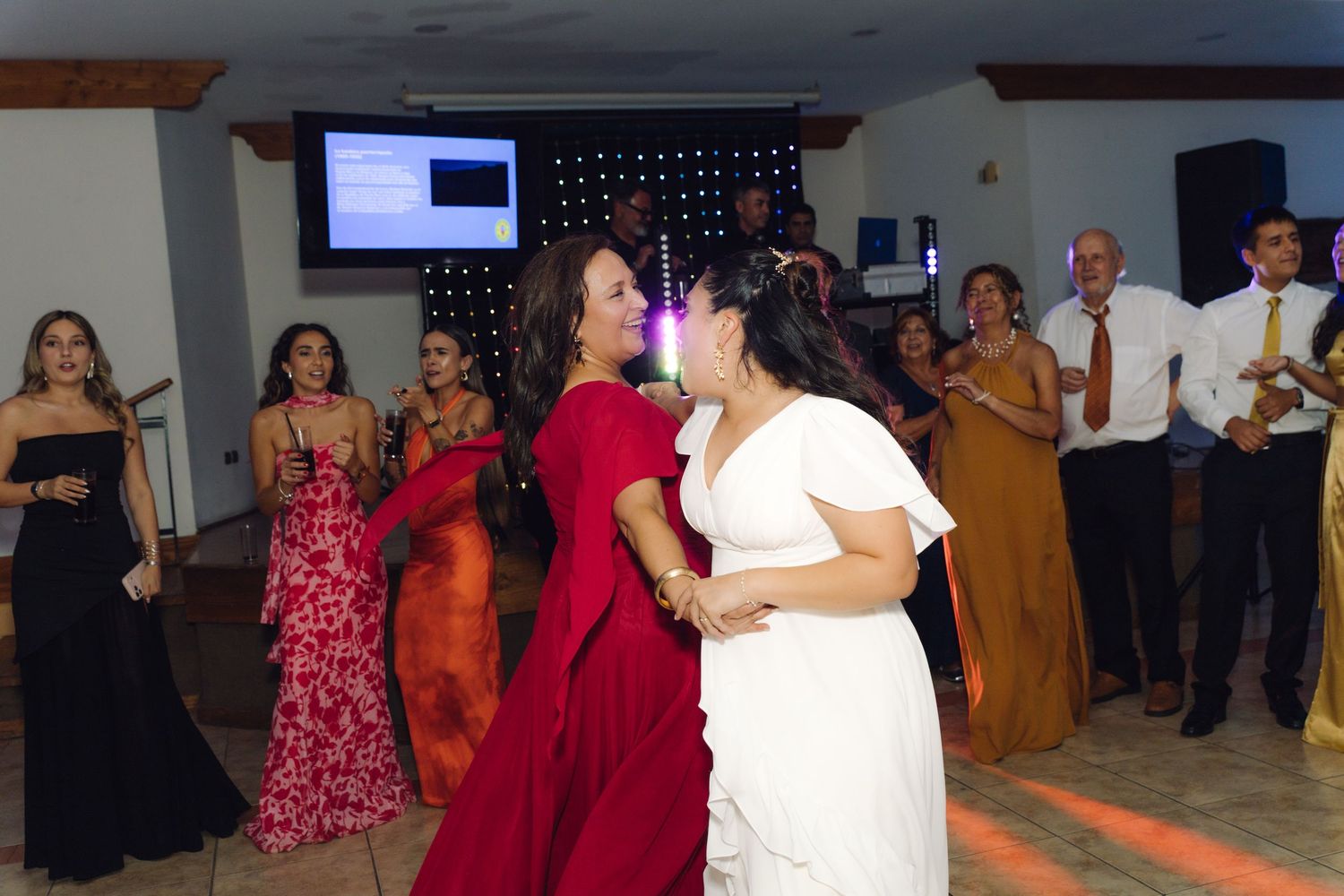 Bride in white dress dances with woman in red gown at a lively wedding reception.